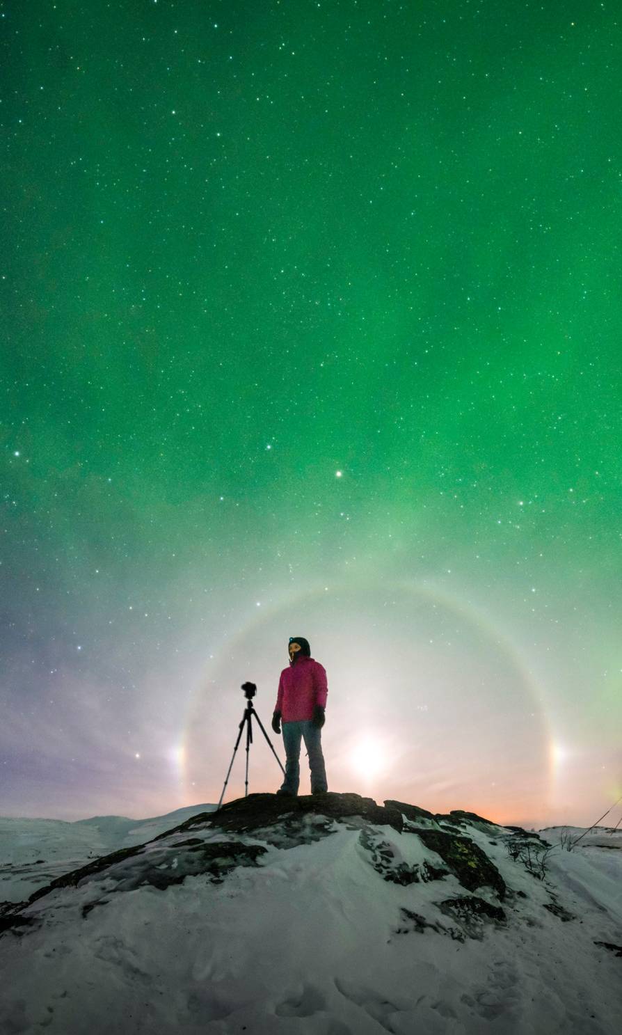 A person standing with its camera on a snowy hill under an epic aurora where the sky is changing colour from blue, yellow, purple, pink and a bright green.