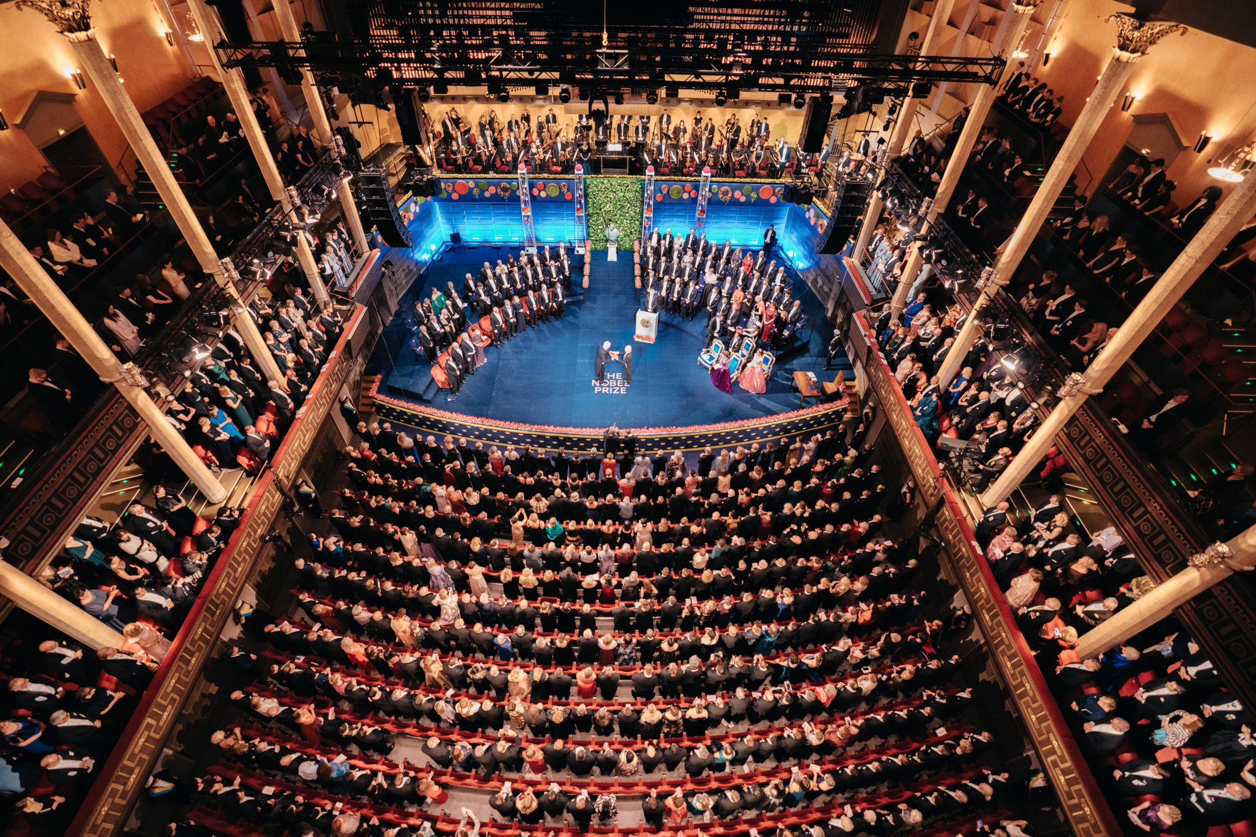 A formal event in a large hall with people seated and standing on stage, dressed in evening attire.