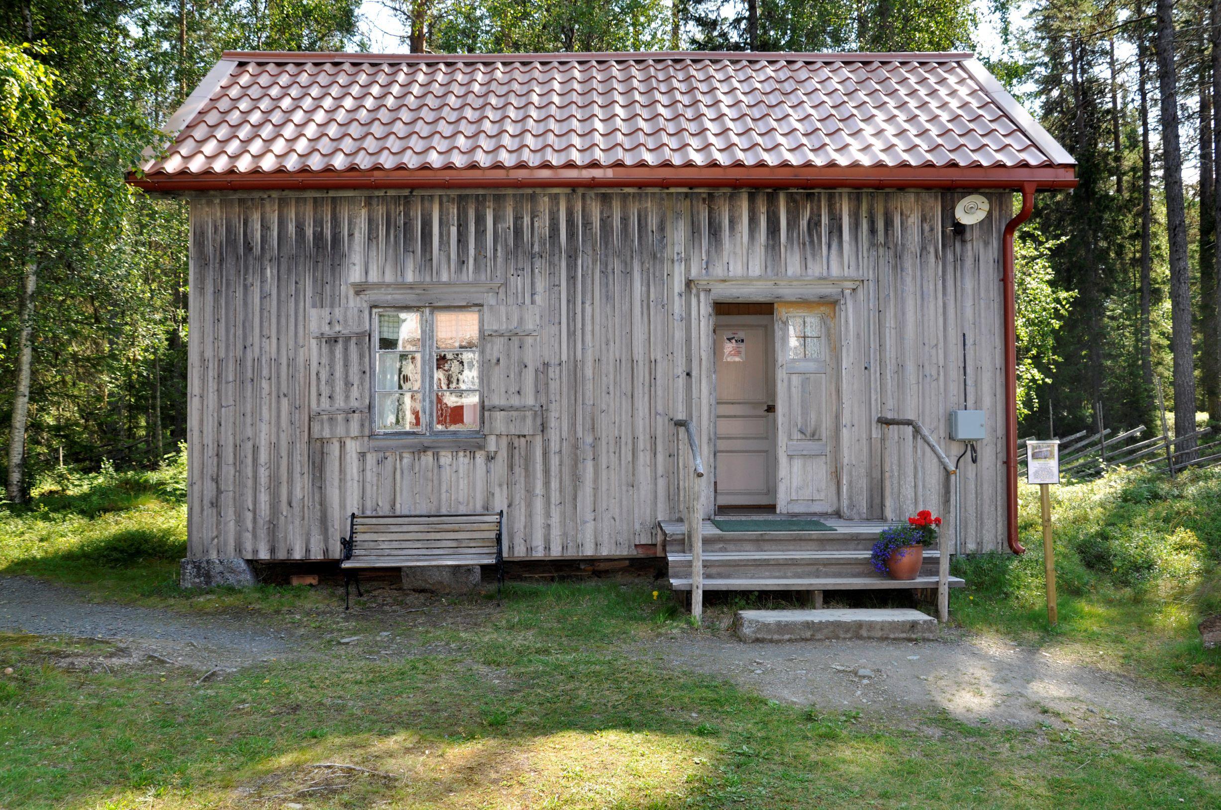 A simple, grey timber cottage with a flowerpot by the entrance, located in front of a forest.