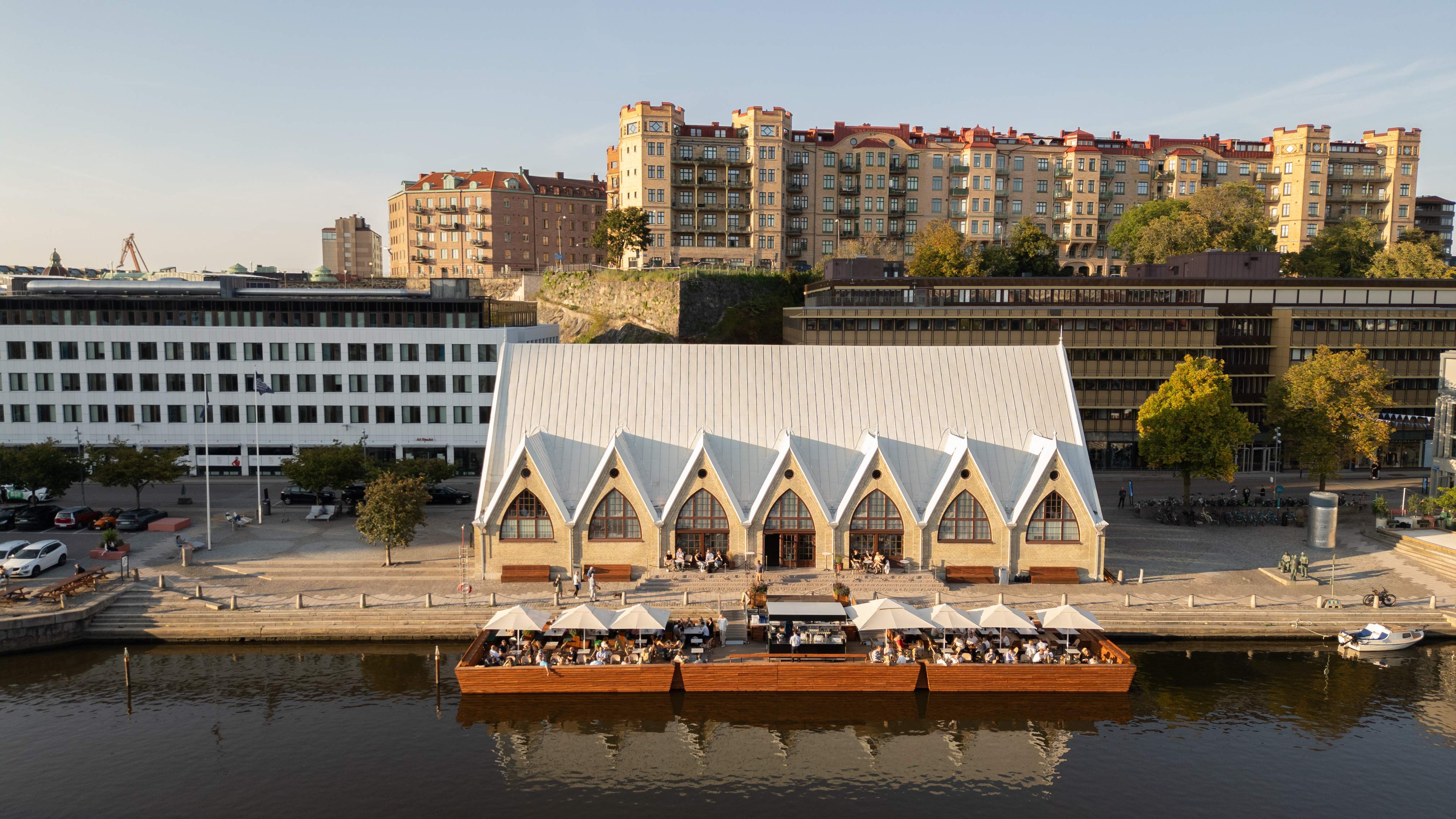 De vismarkt Feskekörka in Göteborg, gezien vanaf de overkant van het water op een zonnige dag, met terrassen en mensen die aan het water zitten te eten.
