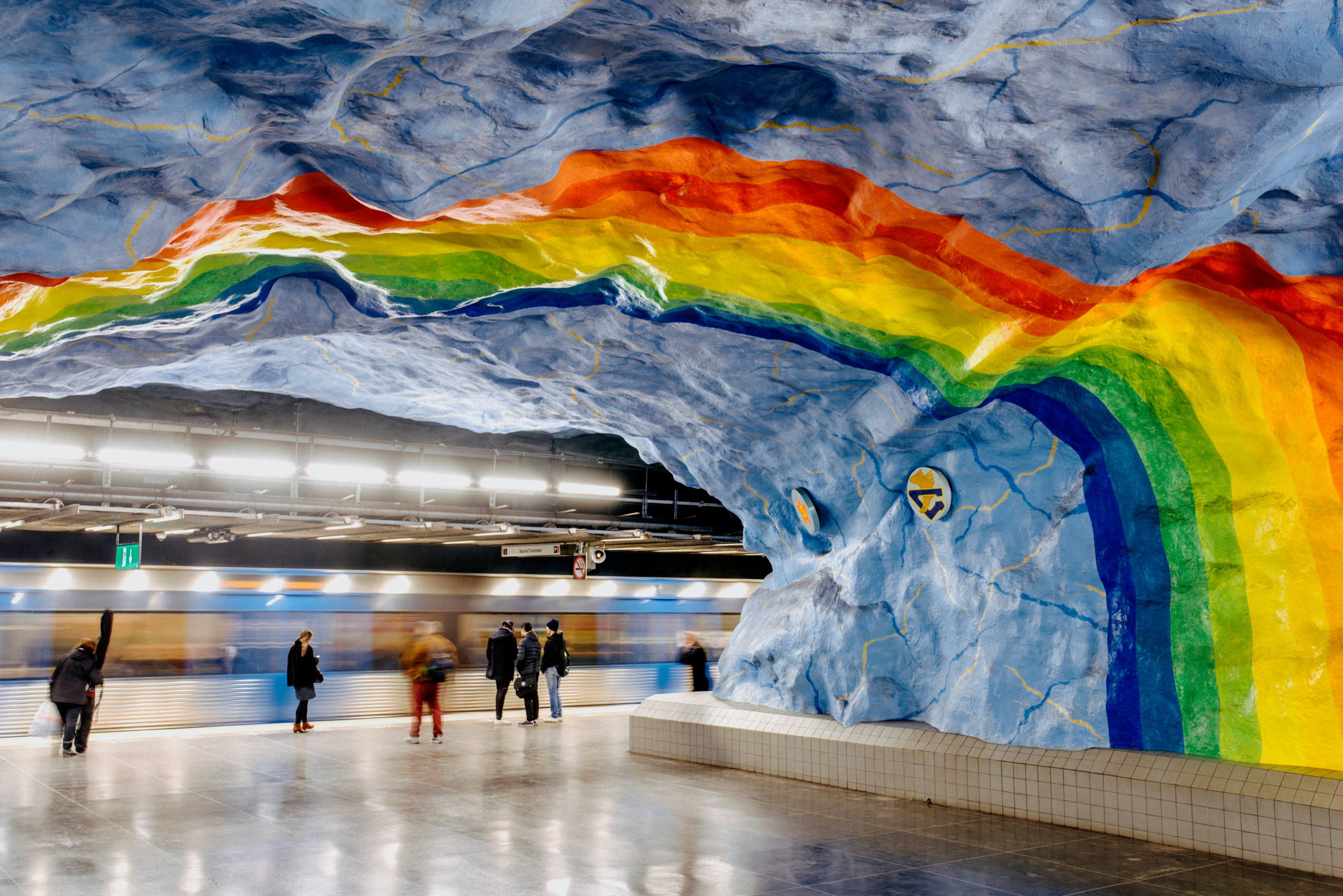 Colourful rainbow mural painted across a rocky ceiling in Stockholm’s metro station Stadion, with people waiting on the platform and a train in motion.