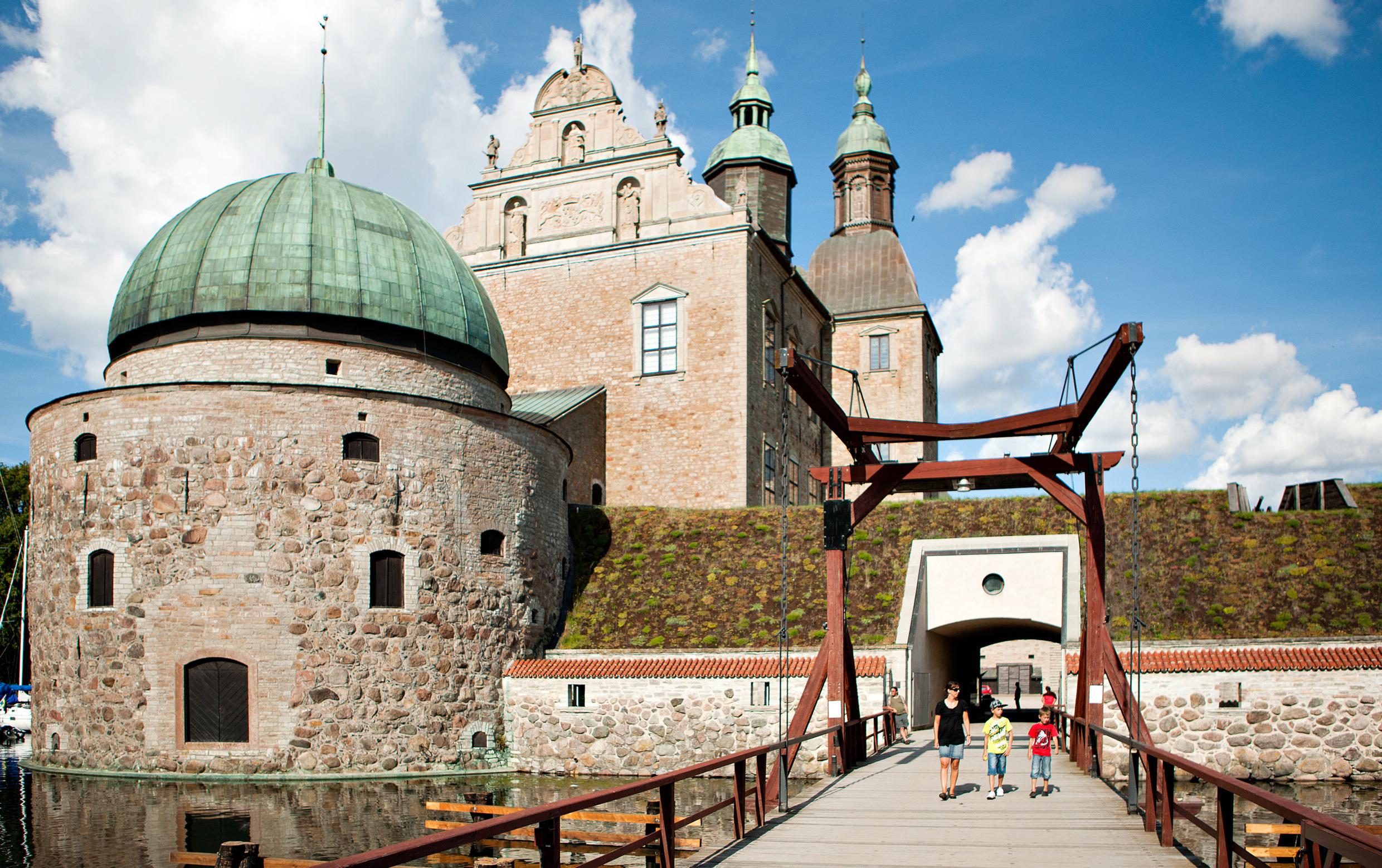 Drie kinderen op een brug die leidt naar het stenen kasteel Vadstena, dat is omgeven door een stenen muur, heg en slotgracht.