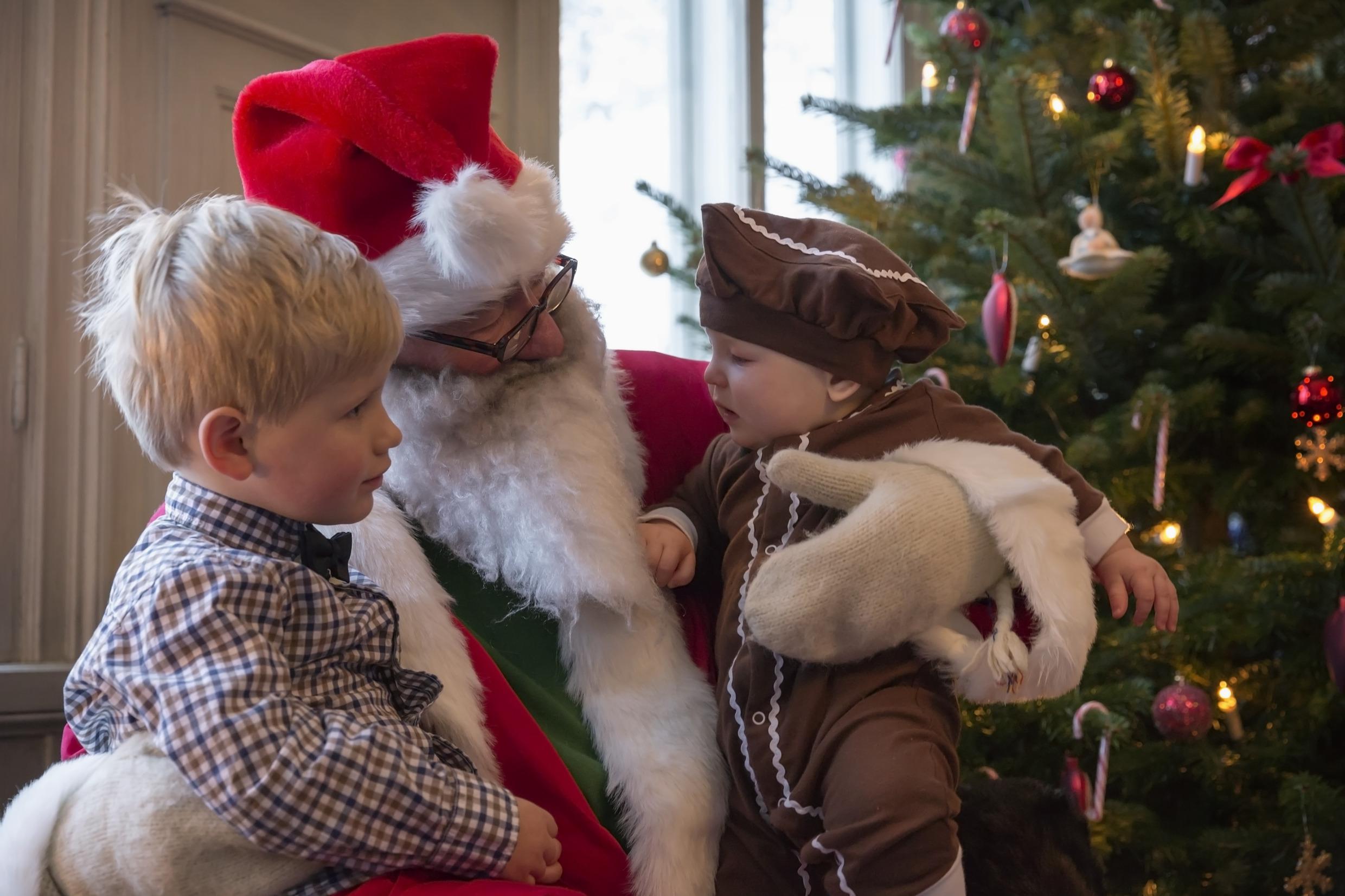 De kerstman zit naast de kerstboom en heeft twee kindjes op de schoot.