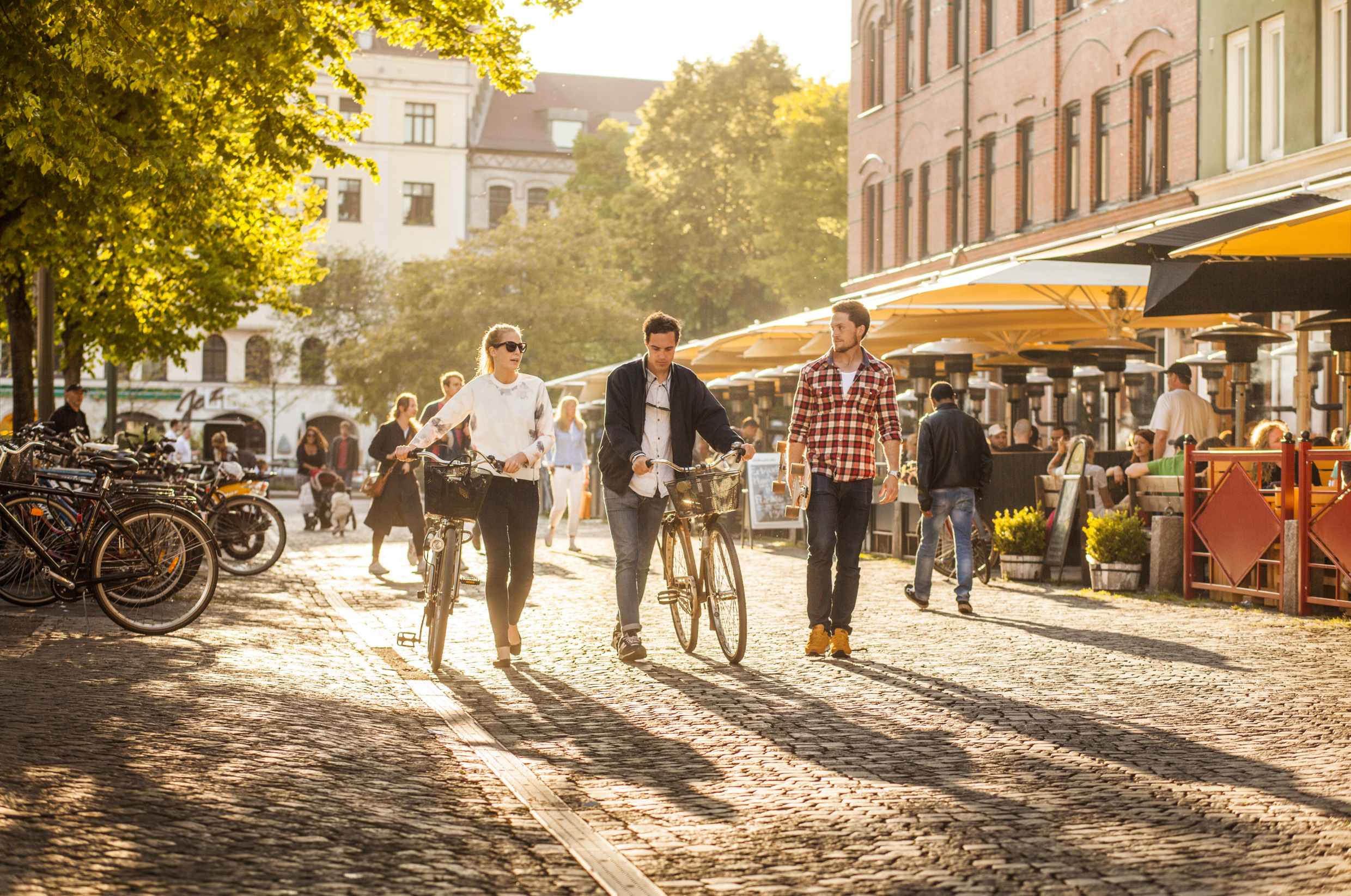 Deux hommes et une femme passent devant des restaurants en plein air sur une place par une journée d'été ensoleillée.