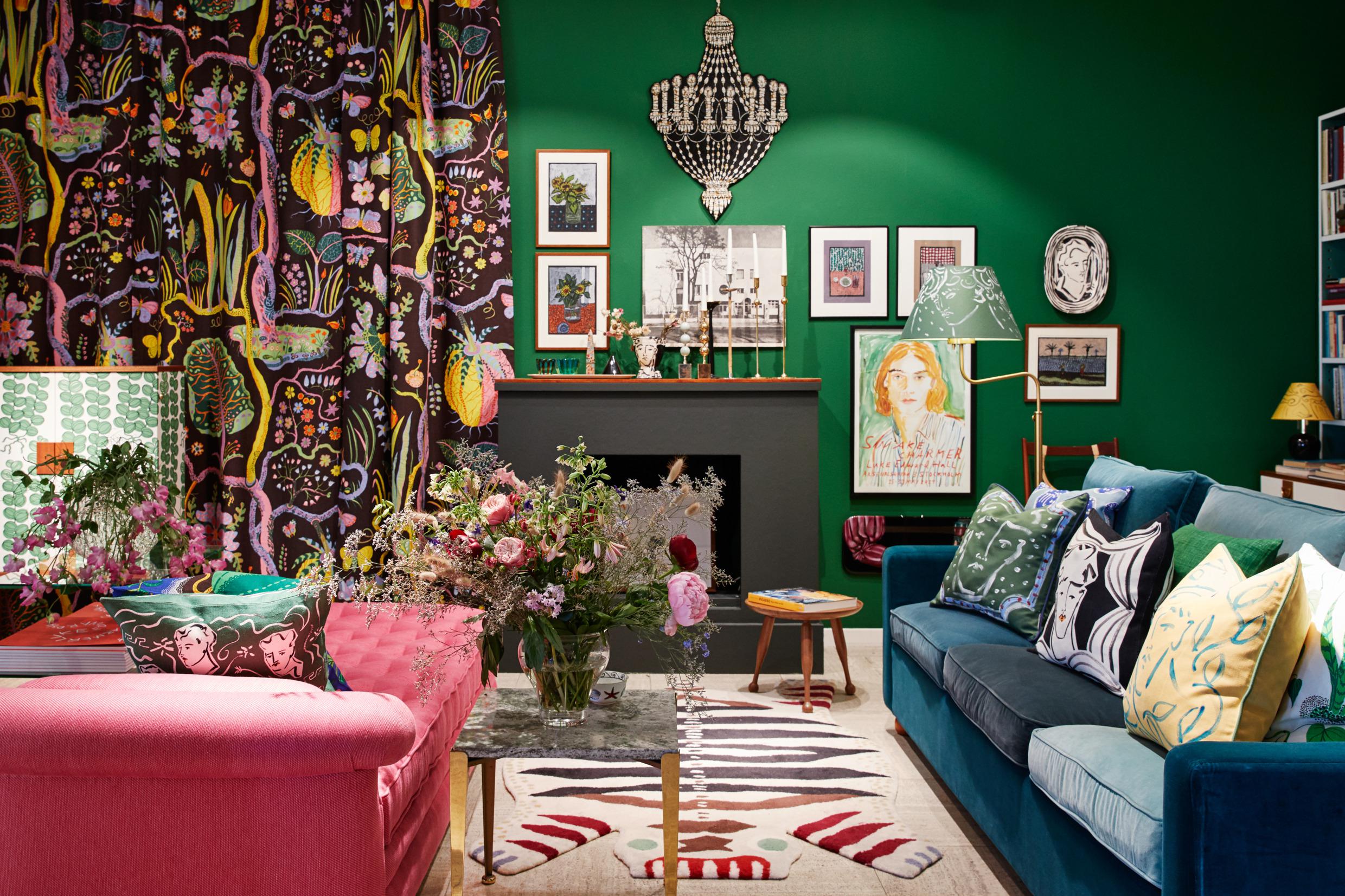 Une chambre colorée avec plusieurs coussins avec des motifs sont posés sur deux canapés. Entre les canapés, il y a un grand bouquet de fleurs posé sur une table. À l'arrière-plan se trouve un rideau à motifs colorés et plusieurs tableaux accrochés sur le mur.