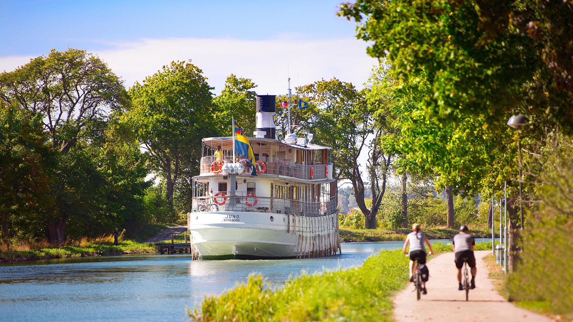 Een zomerse dag. Twee mensen fietsen naast het Göta-kanaal waar een boot nadert.