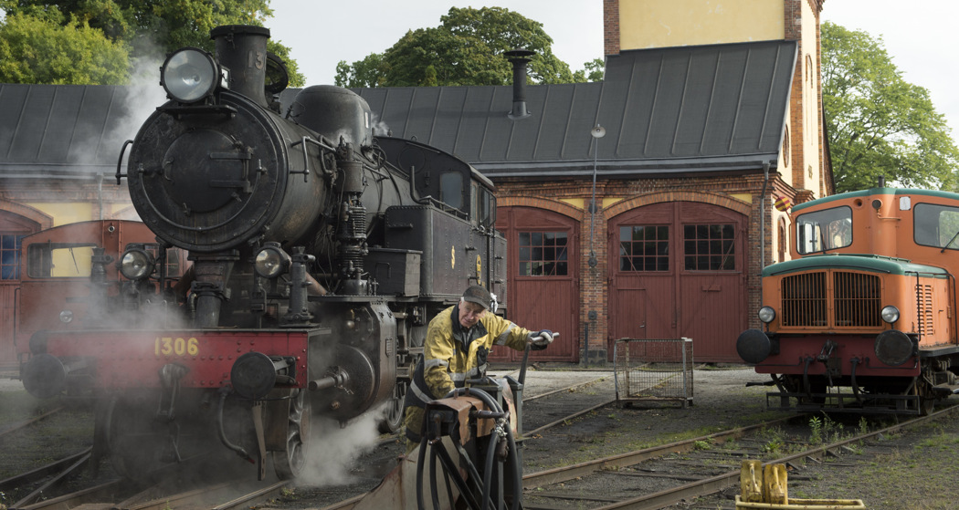 Black steam locomotive being serviced outside a historic brick engine shed in Nora, with a worker and an orange-and-green diesel shunter.