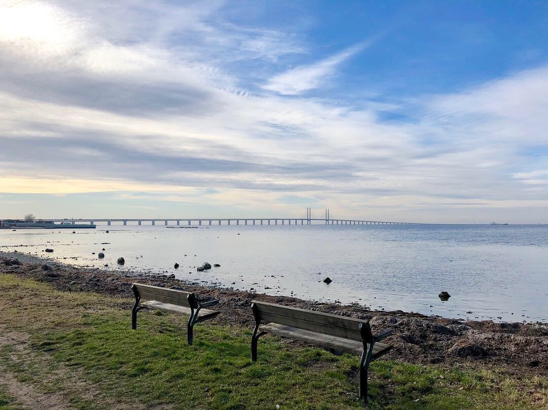 The Öresund Bridge in the background stretching towards the horizon. The sea is calm and the park benches in front of the waterline are empty.
