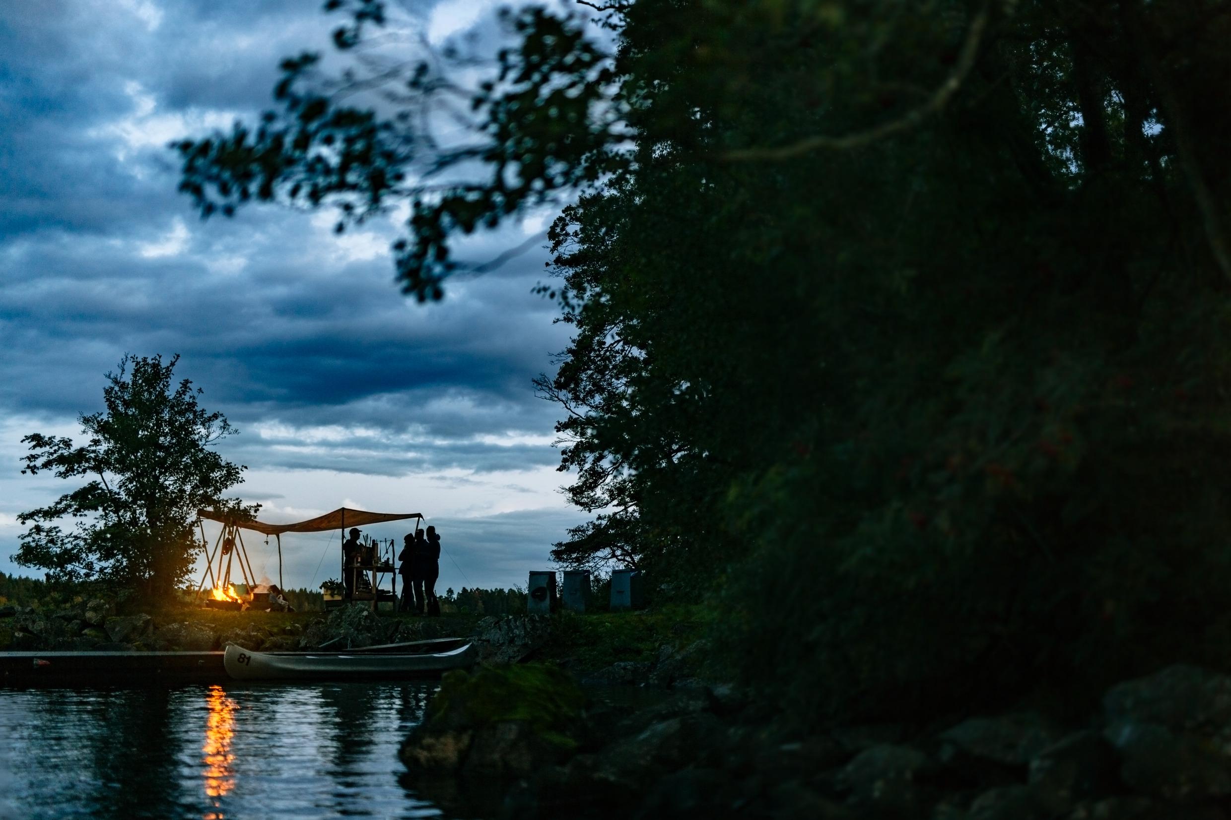 A group of people in Sweden are standing by a campsite and camp fire in the evening, next to a lake with a canoe.