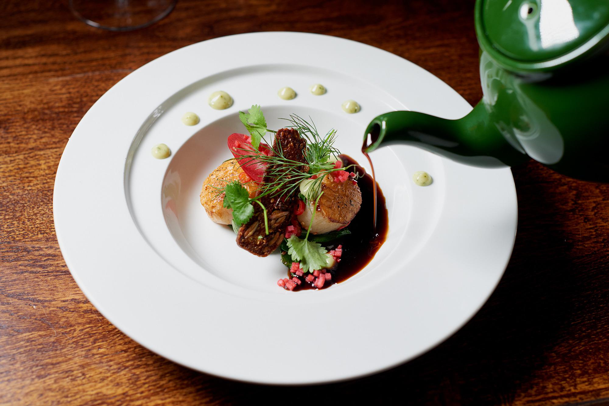 A dish of scallops and locally sourced ingredients från Österlen at restaurant Hickory's Bistro in Simrishamn. A brow sauce is being poured on the plate from a green porcelain jug.