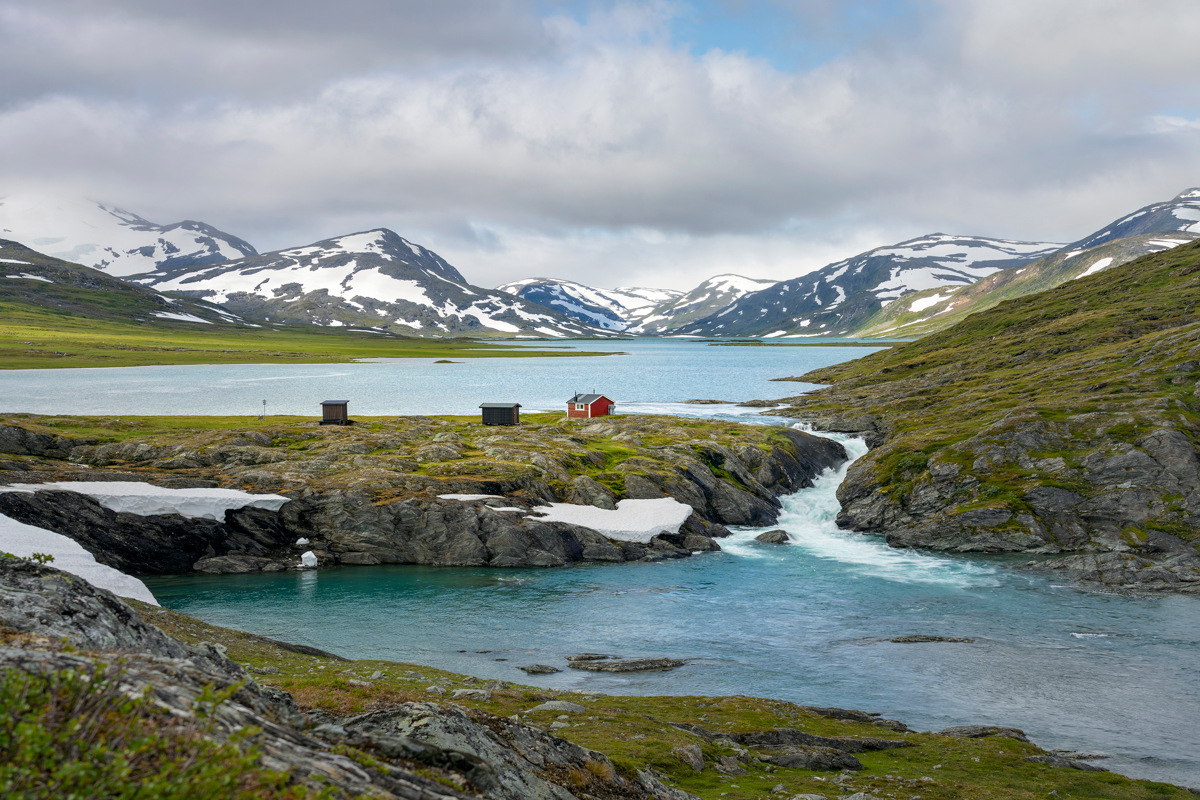 Berglandschaft mit dem See Sårjåsjávrre und der kleinen roten Hütte von Konsul Persson, umgeben von schneebedeckten Gipfeln.