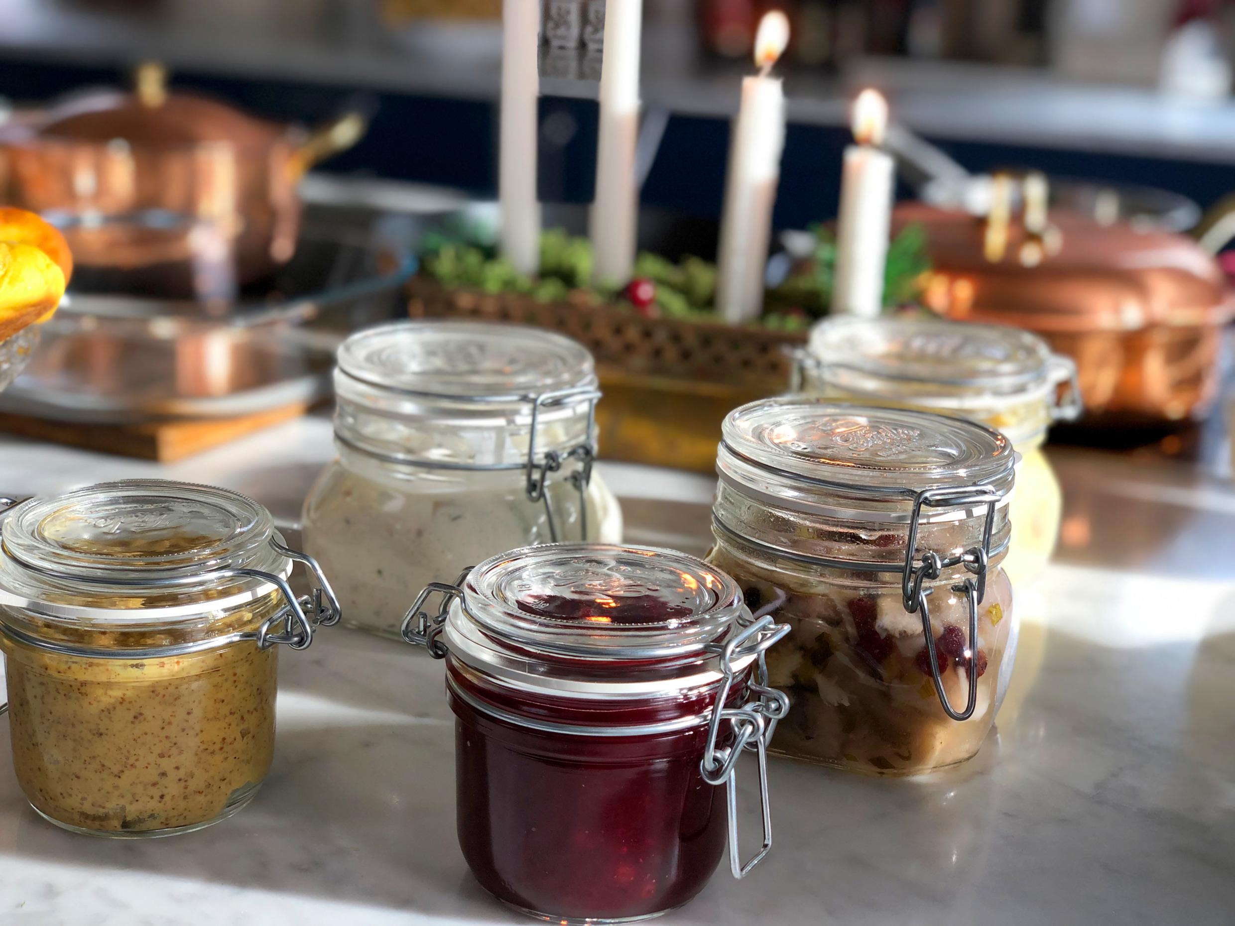 Close-up van vijf glazen potten met ingemaakte haring, adventskandelaar met brandende kaarsen en een koperen pot op de achtergrond.