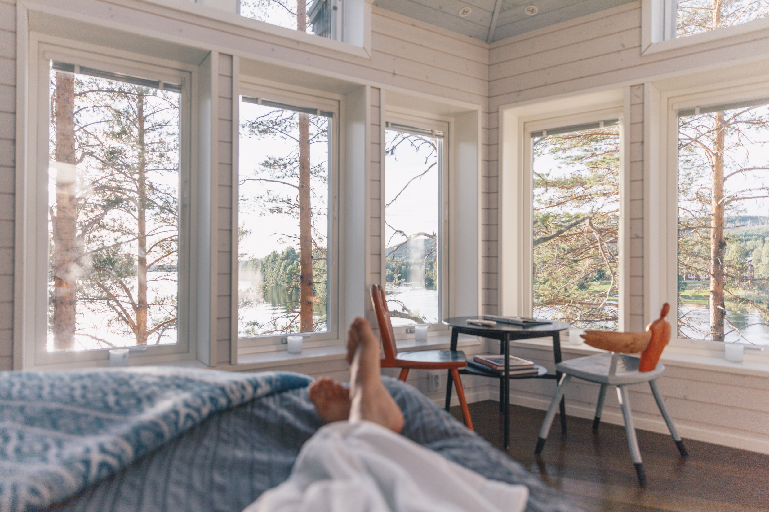 Die Beine von jemandem, der auf einem Bett in einem Baumhaus liegt. Es gibt mehrere Fenster und einen tollen Blick auf einen Fluss und die Natur draußen.