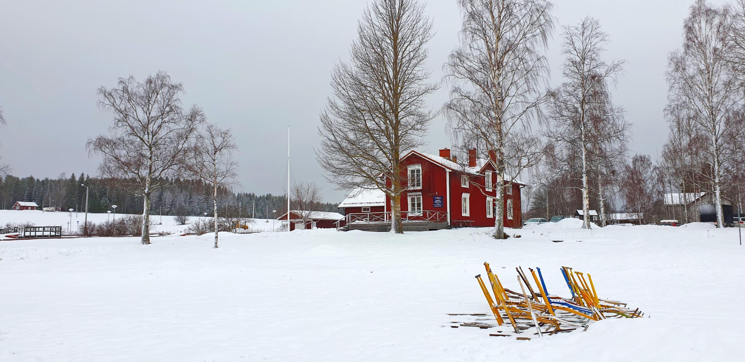 Ein rotes Haus im Winter. Vor dem Haus steht eine Reihe von Tretschlitten.
