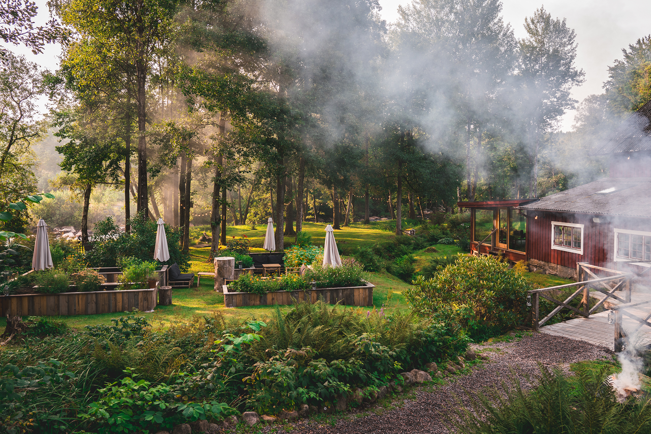 A restaurant setting in a green forest with outdoor seating, wooden buildings and sunlight filtering through the trees.