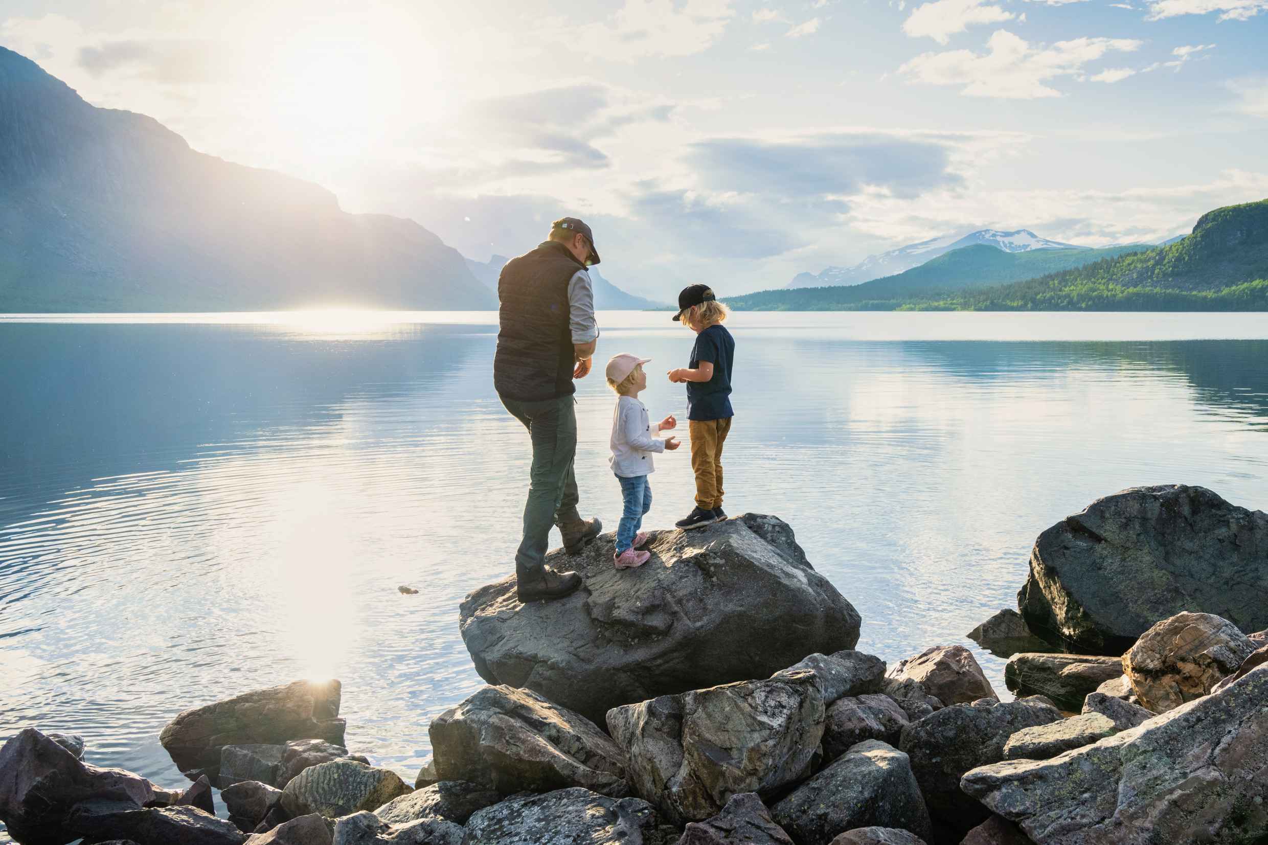 Een volwassene en twee kinderen staan op rotsen bij een stil bergmeer, met zacht avondlicht en dramatische bergtoppen op de achtergrond.