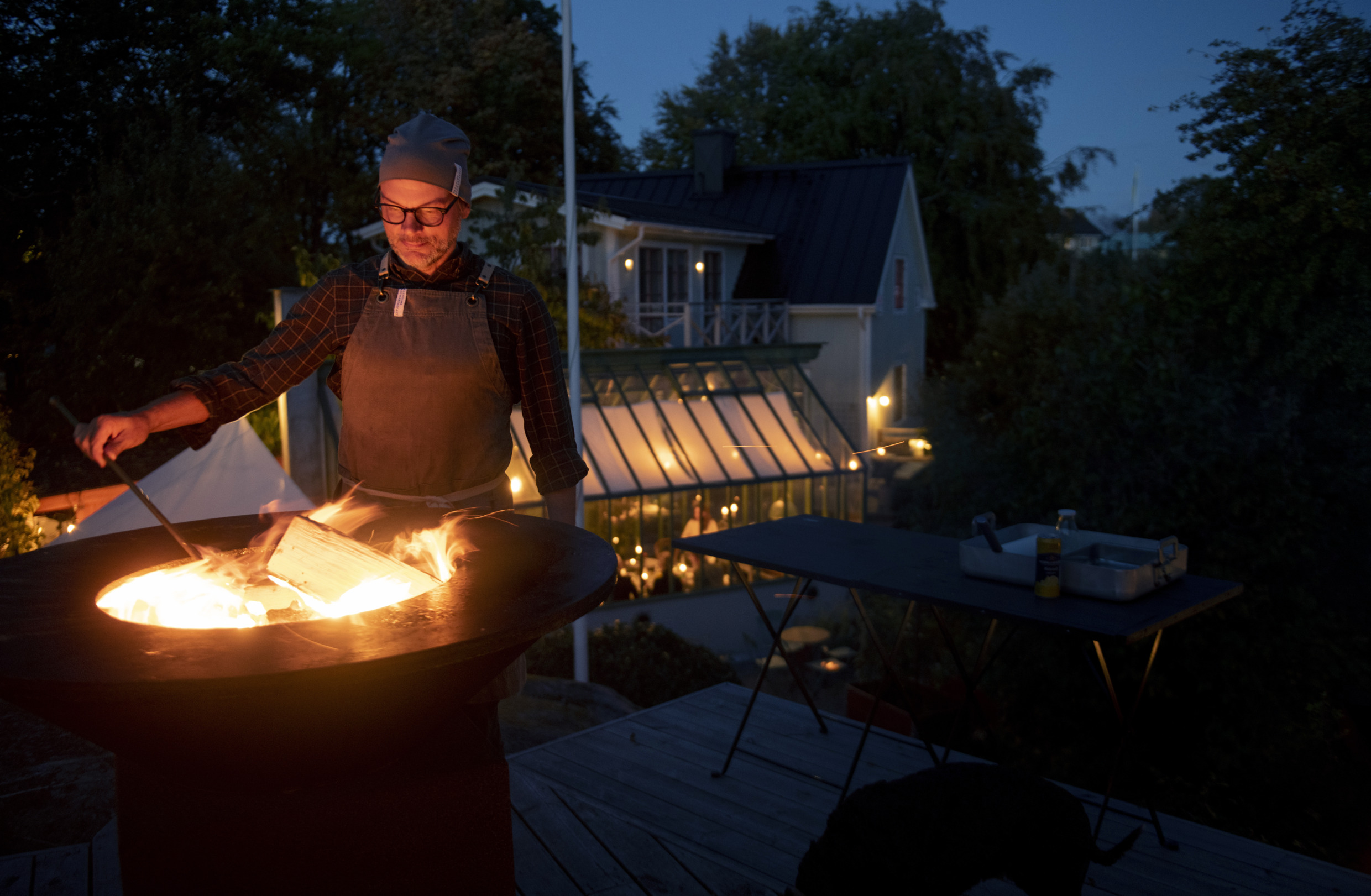 A man with a leather apron is standing by a fire during the evening.