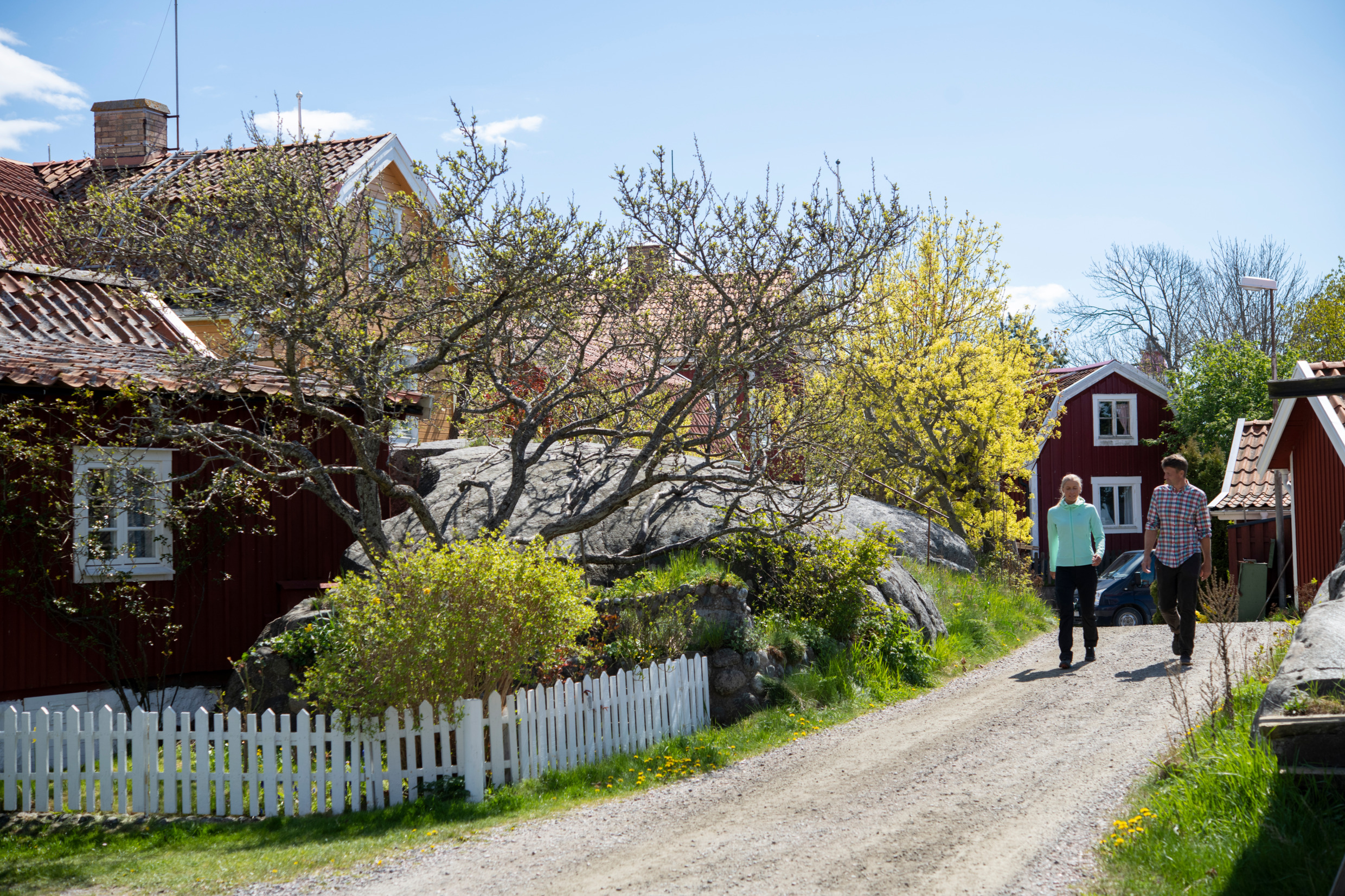 A man and a woman walking on a road next to red wooden houses and white wooden fences.