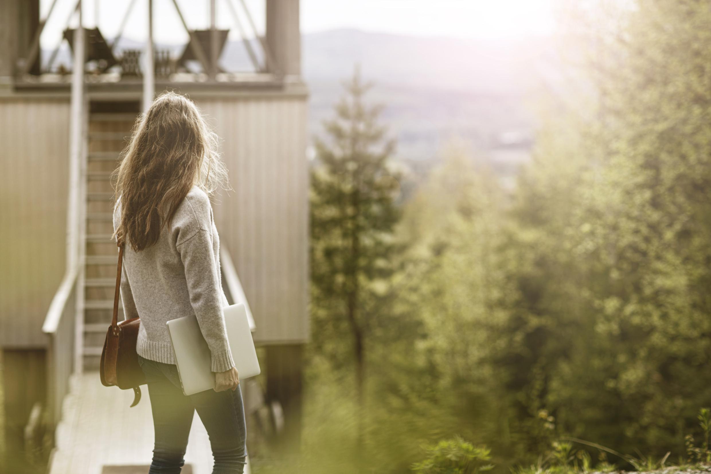 Een vrouw met een laptop kijkt naar een houten huis met grote ramen op een heuvel in de natuur.