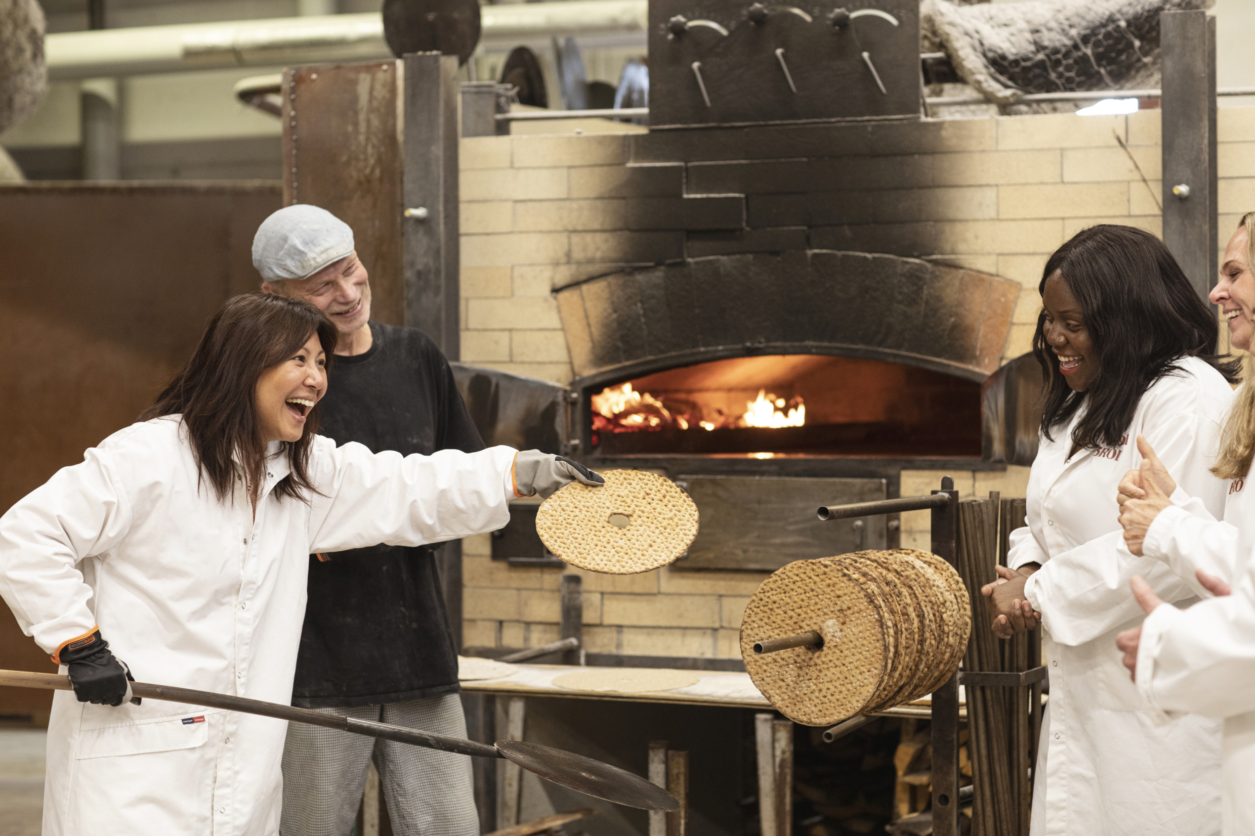 Drei Frauen und ein männlicher Guide stehen vor Holzöfen in einer Bäckerei. Eine der Frauen zeigt den anderen ein frisch gebackenes Knäckebrot.