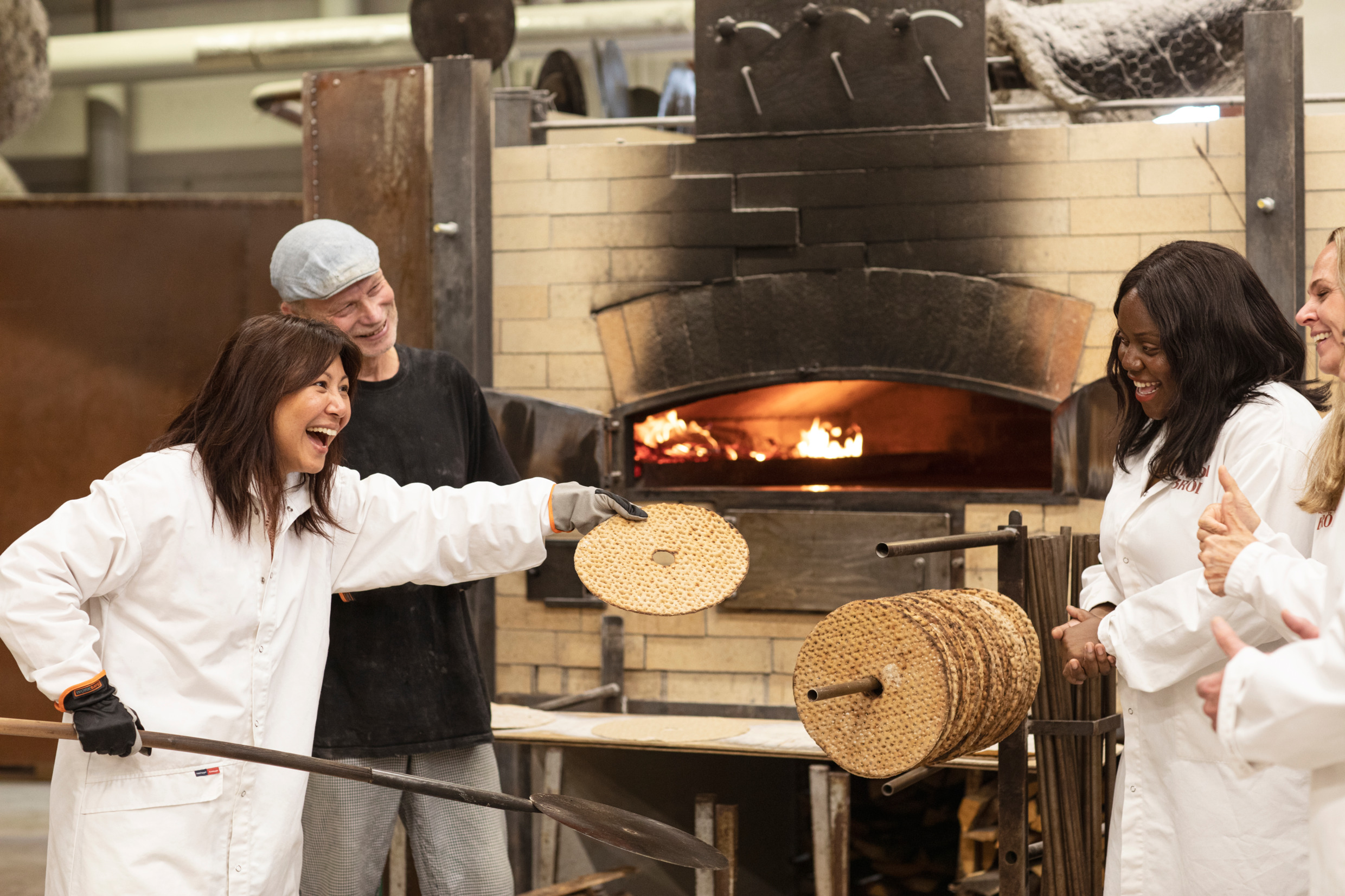 Three women and a male guide are standing in front of wood fired ovens in a bakery. One of the women are showing a freshly baked crispbread to the others.