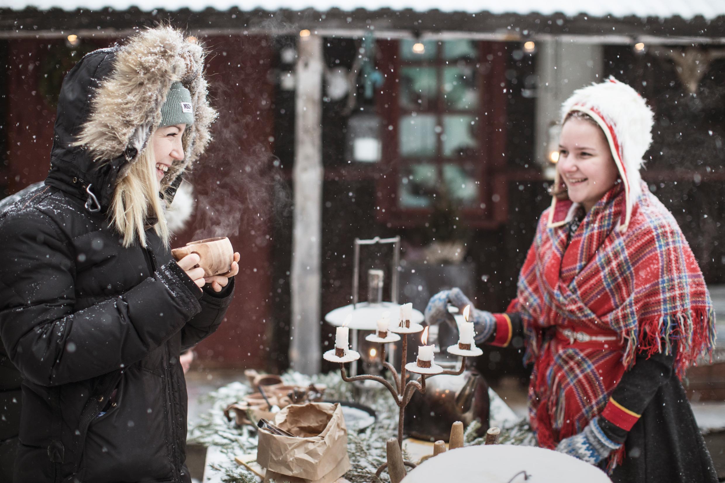Zwei Frauen stehen an einem Tisch und halten eine Fika im Freien im Schnee. Auf dem Tisch steht ein alter Kandelaber mit brennenden Kerzen. Eine der beiden Frauen trägt eine samische Tracht. Es schneit.