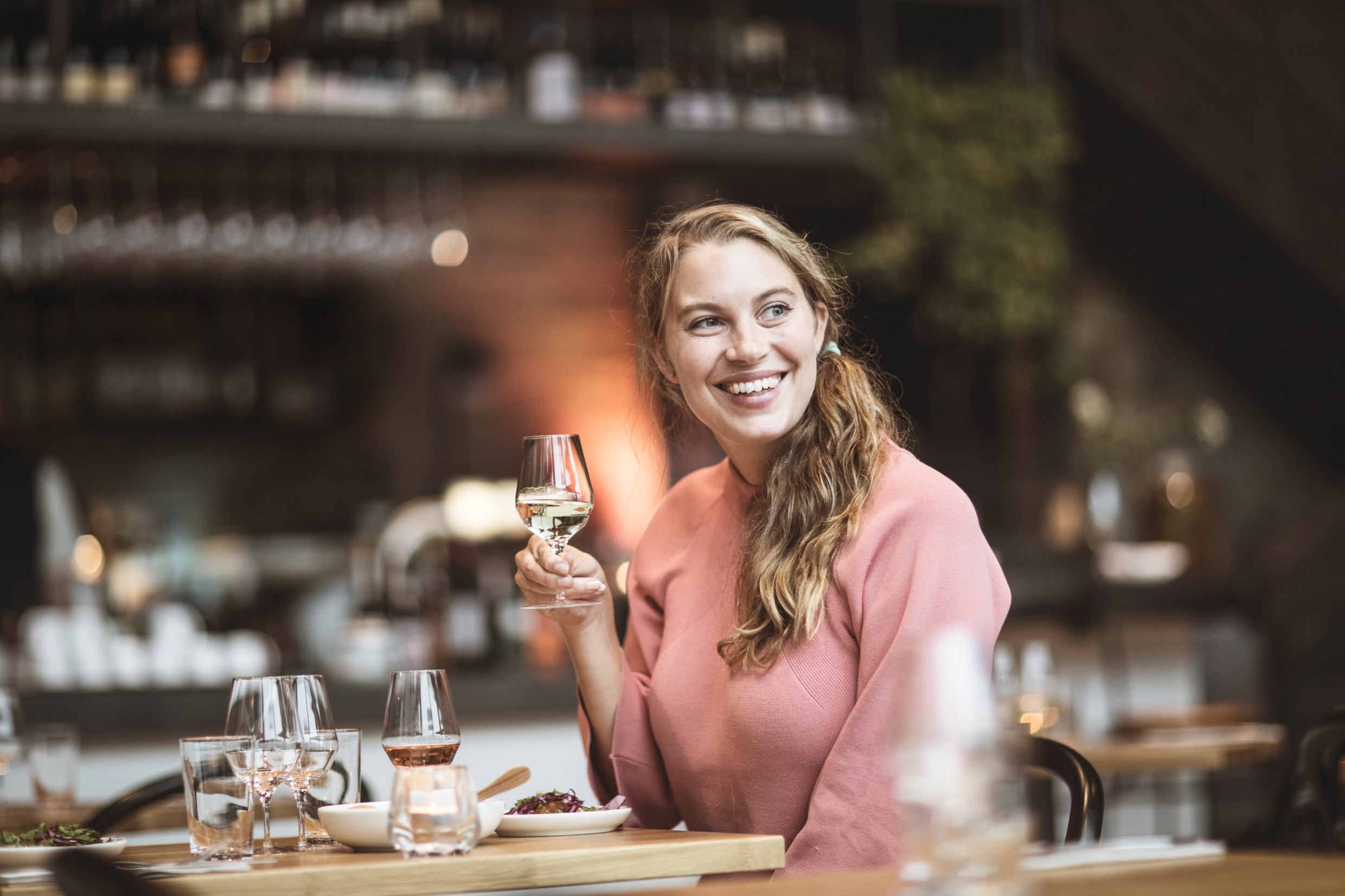 Eine lächelnde Frau, die an einem Tisch in der Weinbar Wine Mechanics in Göteborg sitzt und ein Weinglas in die Höhe hält, als würde sie jubeln.