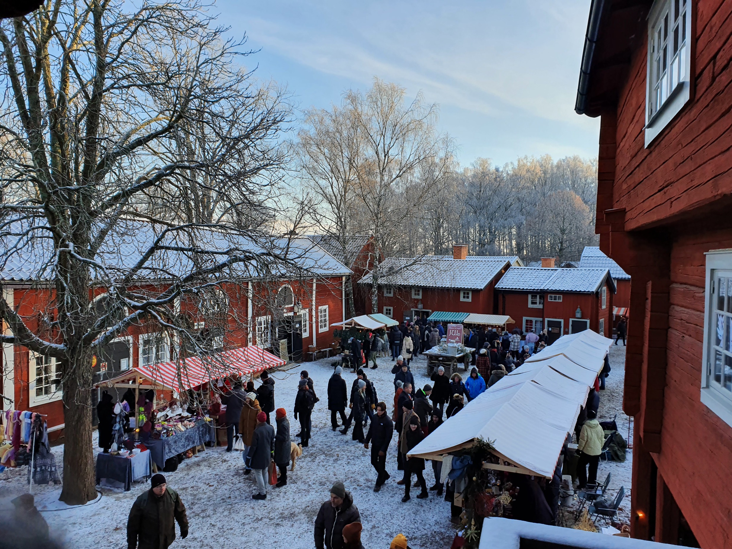 Des visiteurs explorant les étals recouverts de neige parmi les maisons en bois rouges du musée en plein air de Gamla Linköping.