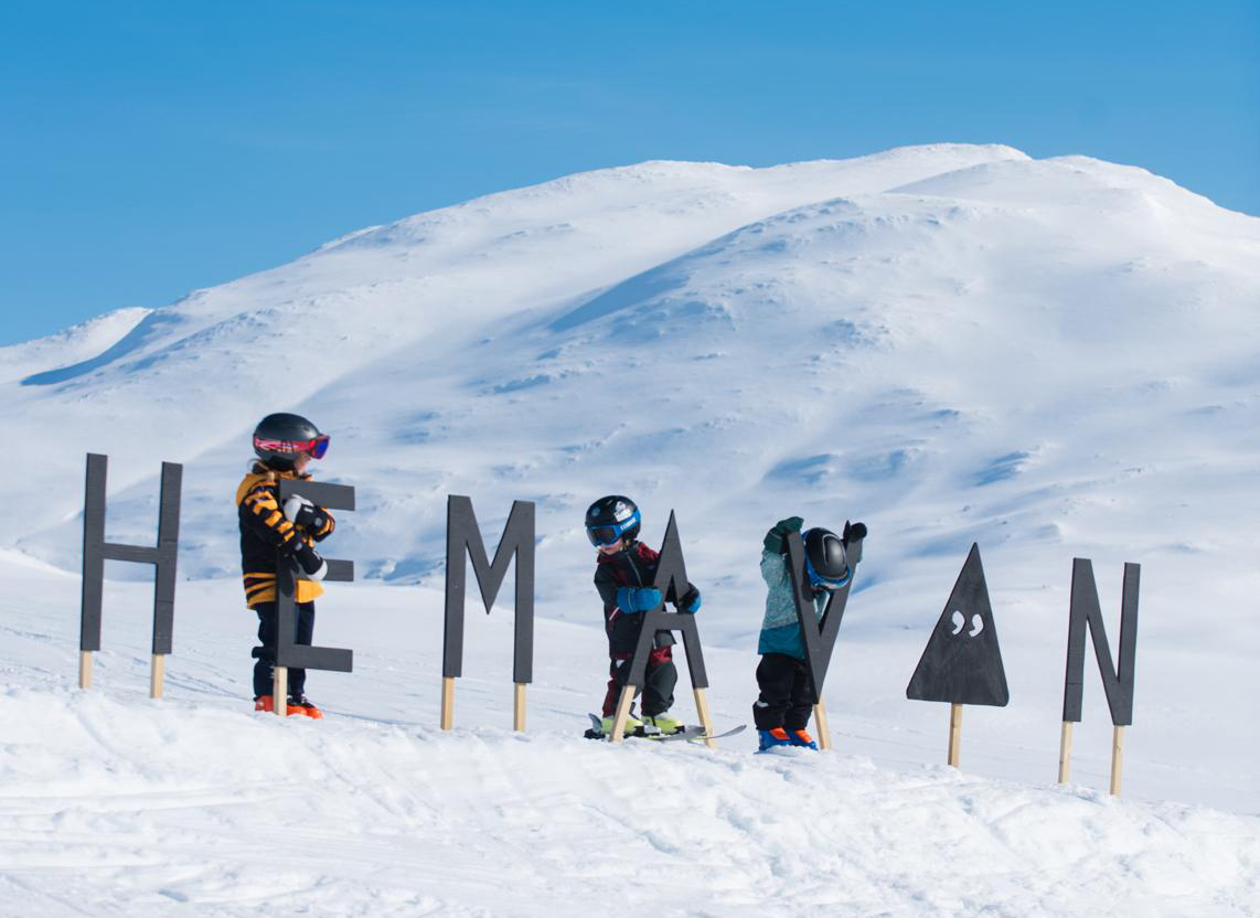 Three children in ski gear playing beside large "Hemavan" letters with snowy mountains in the background.