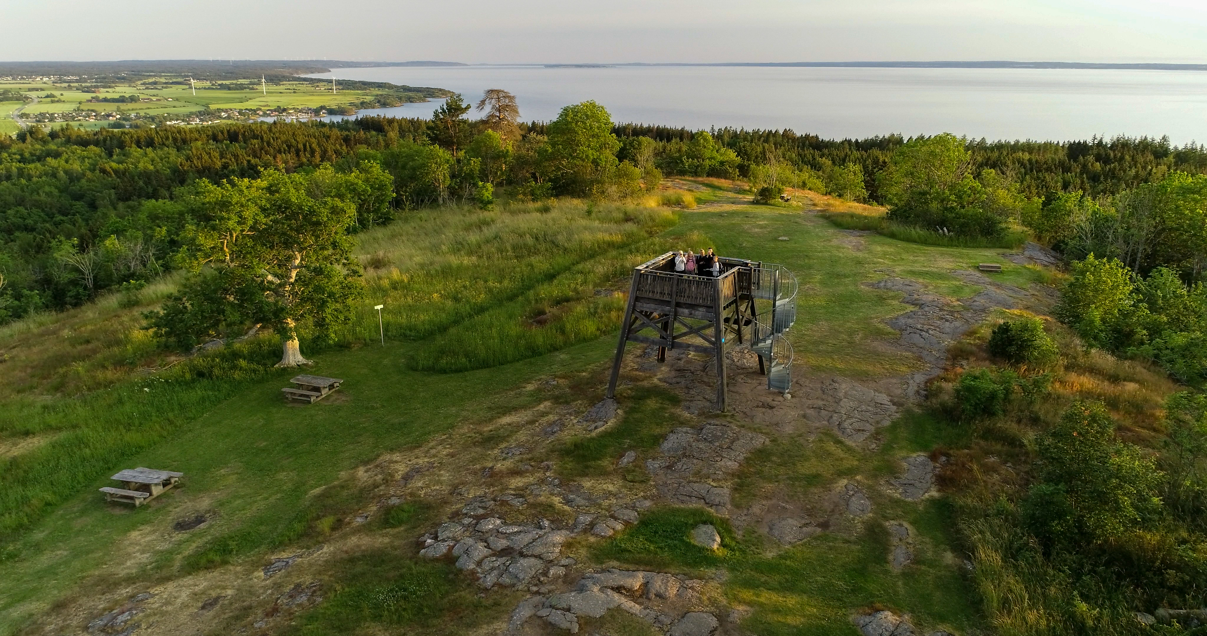Viewpoint tower at Hjässan on Omberg with people standing on it, surrounded by grassy landscape, trees and a view of Lake Vättern.