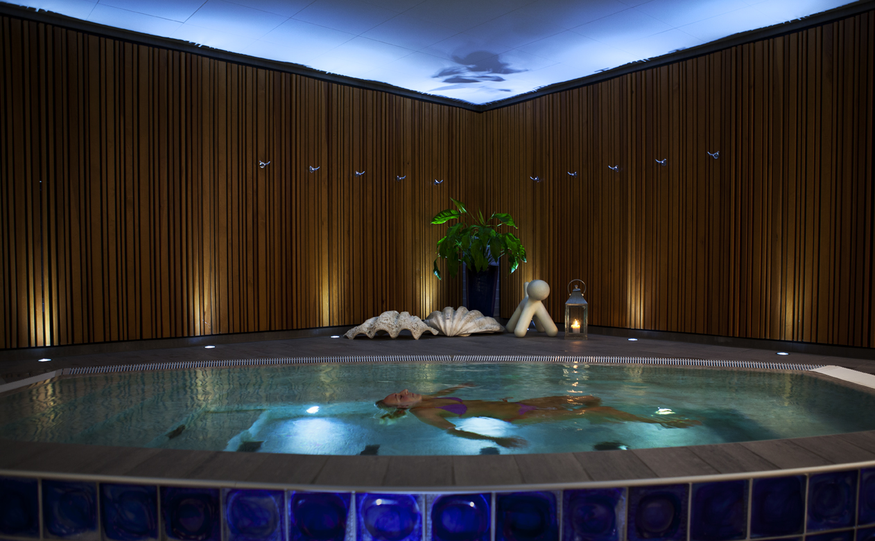 Un client flottant dans la piscine intérieure chauffée du spa du Kosta Boda Art Hotel, entouré de murs en bois, d'un éclairage tamisé et de détails inspirés du verre.
