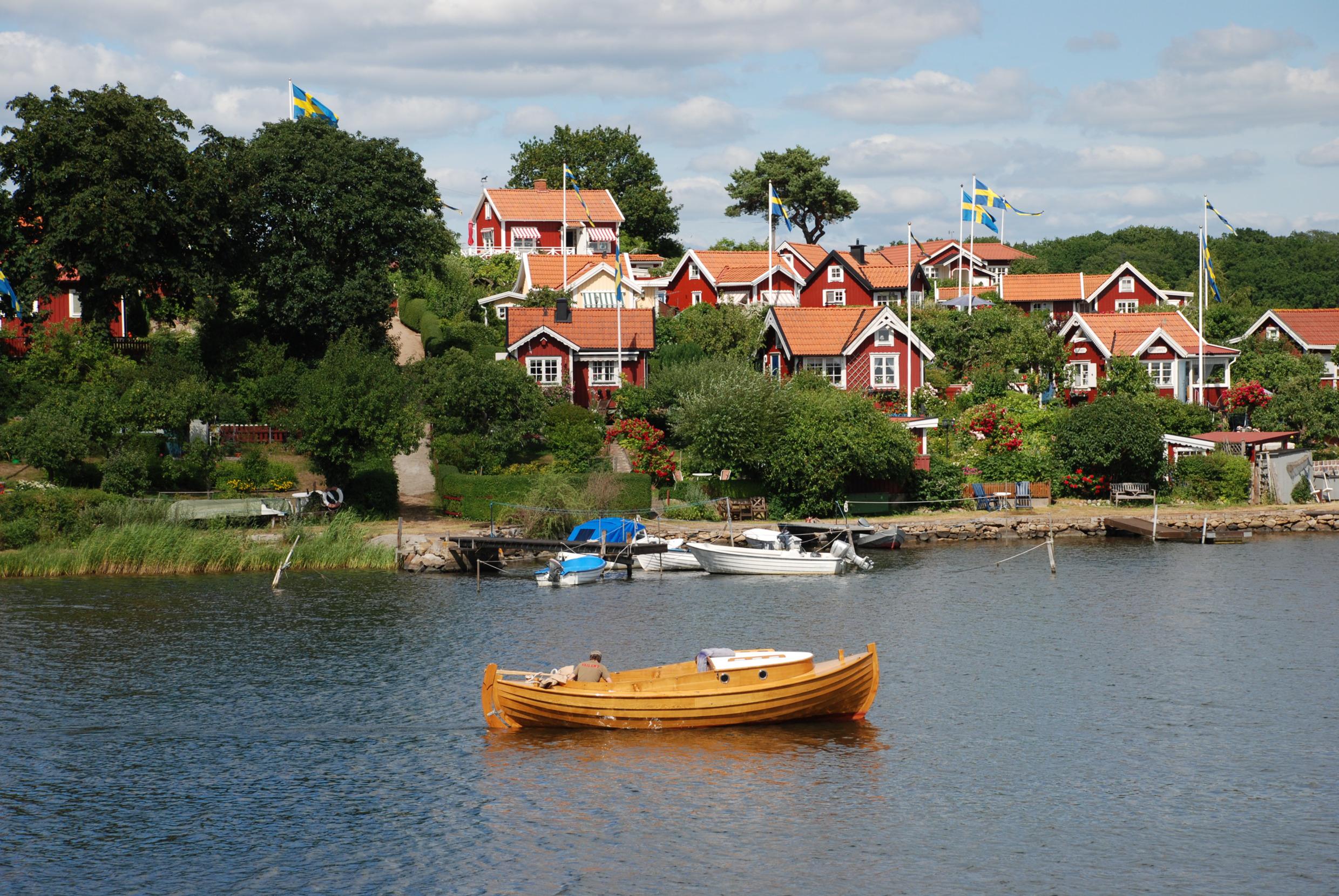 Several small red cottages with the Swedish flag hoisted, nestled in greenery by the sea. A wooden boat is passing by at sea.