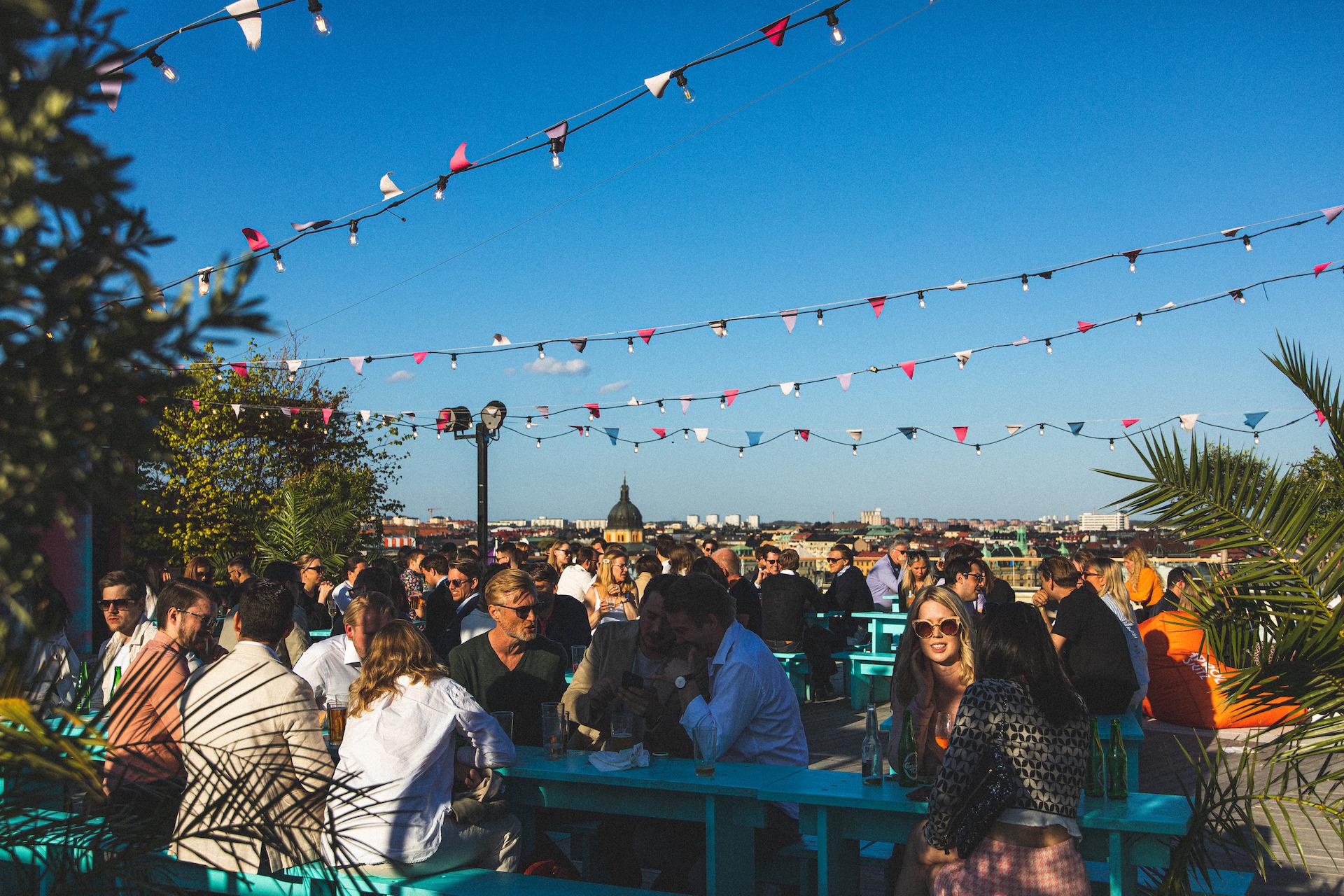 Mensen zitten aan tafels op het dak van Stockholm onder Stjärnorna onder een blauwe lucht. Achter hen is een uitzicht op Stockholm.