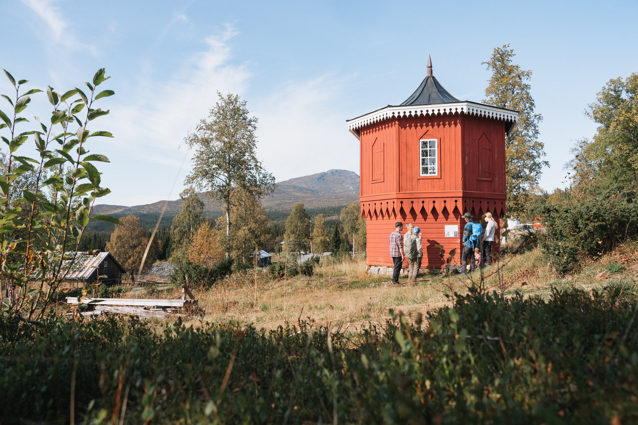 Eine Gruppe Leute steht vor einem roten Holzgebäude in Fröå Gruva, mit herbstlichen Bäumen und Bergen im Hintergrund.