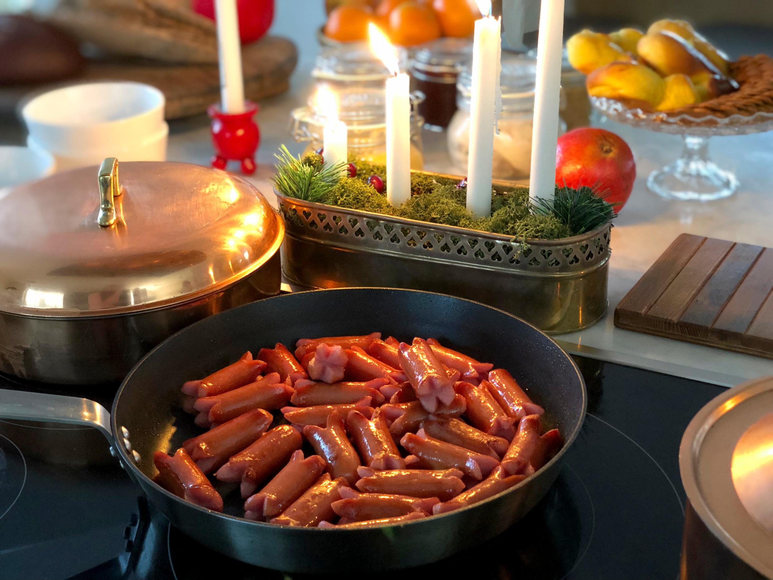 Close up van een koekenpan met prinsenworstjes naast een adventskandelaar met aangestoken kaarsen en een koperen pot. Vruchten op de achtergrond.