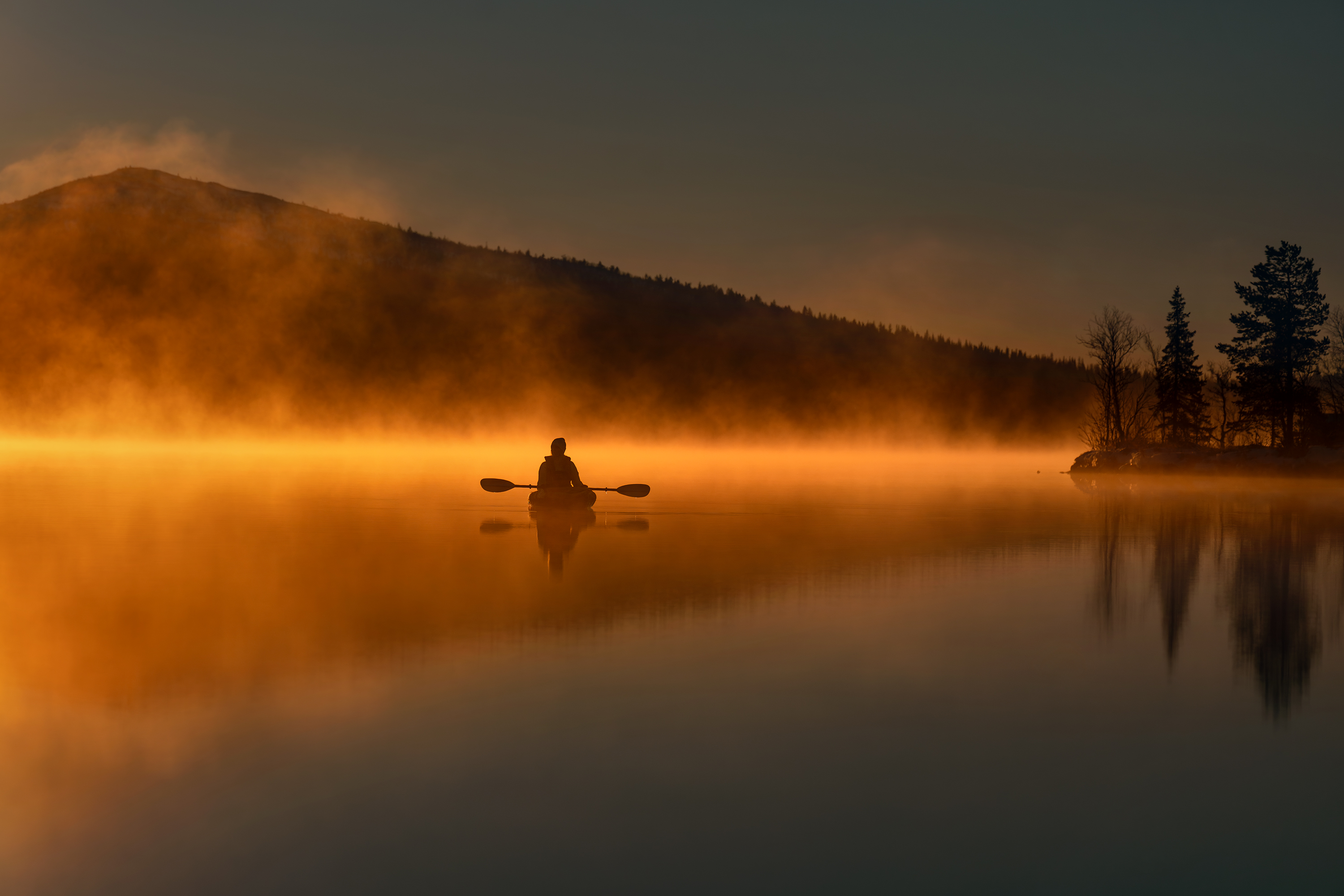 Silhouette eines Kajakfahrers, der bei Sonnenaufgang in Gällivare, im arktischen Schweden, über einen nebligen See paddelt, umgeben von herbstlich gefärbten Hügeln.
