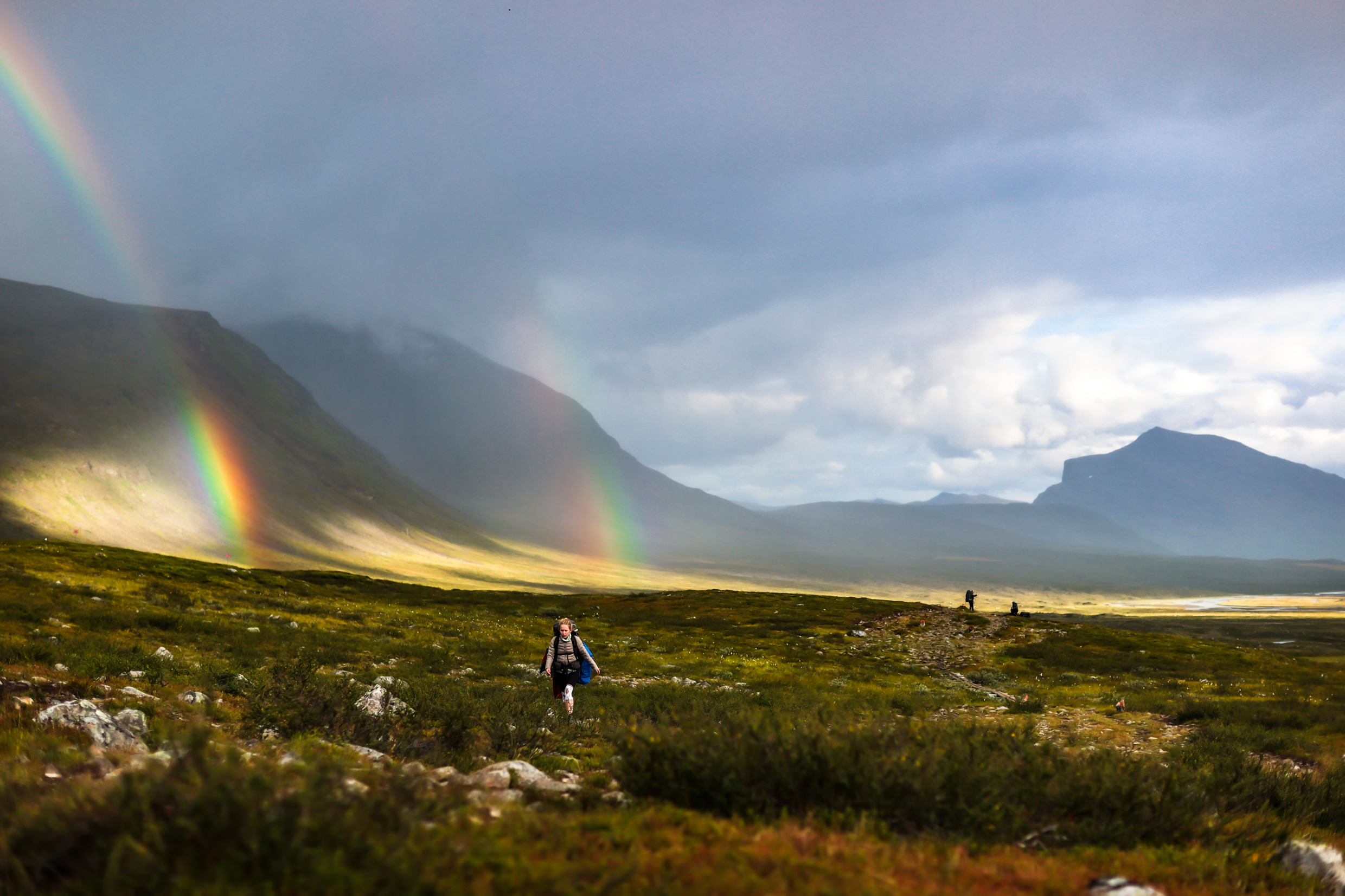 A woman hiking Kungsleden. A majestic view of the mountains, fields and two rainbows.