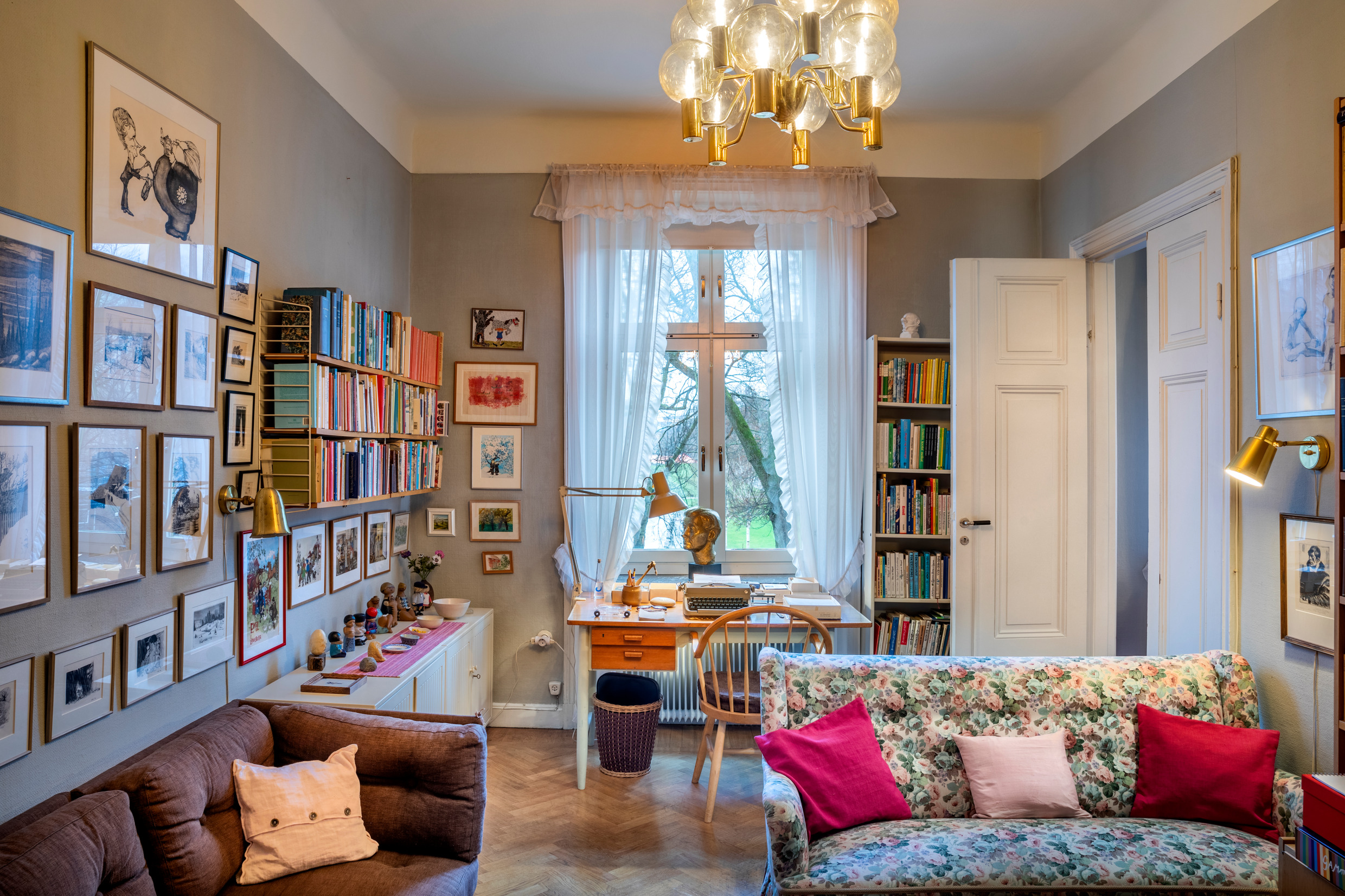The interior of a room tightly filled with a desk, sofas, bookshelves and art on the walls.