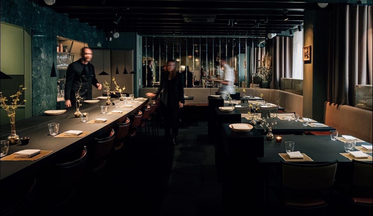 Eine Sitzecke in einem Restaurant am Abend. Drei verschwommene Personen decken die Tische ein.