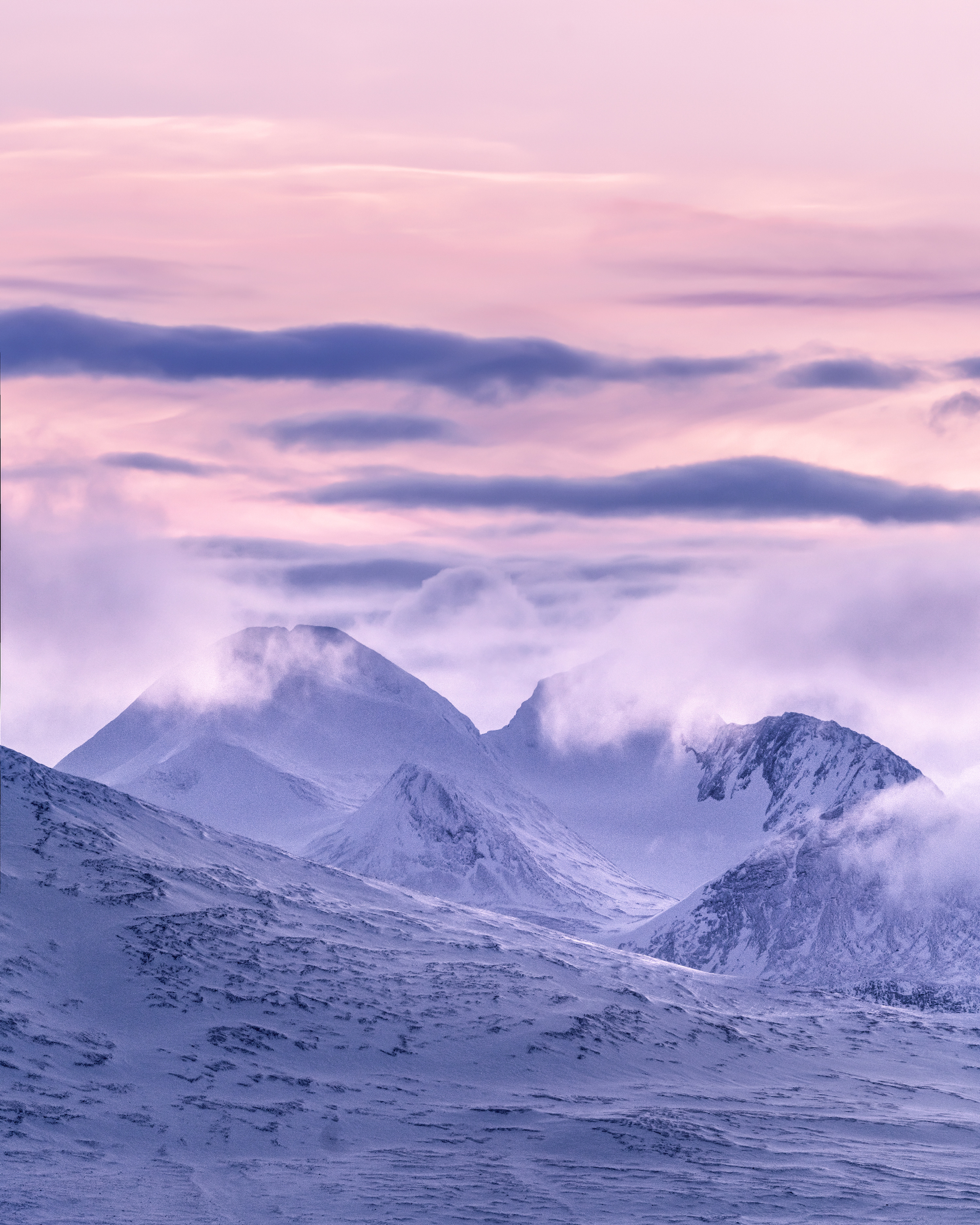 Die schneebedeckten Berge werden teilweise von Wolken verhüllt, die im arktischen Winterlicht rosa leuchten.