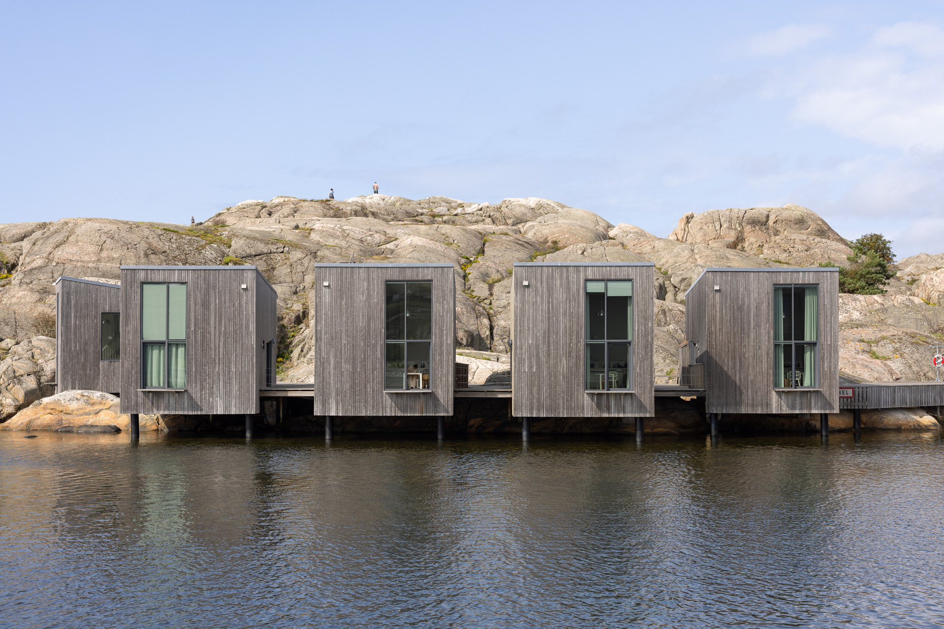 The Nordic Watercolour Museum’s dark wooden buildings by the water, with rocky cliffs rising behind them.