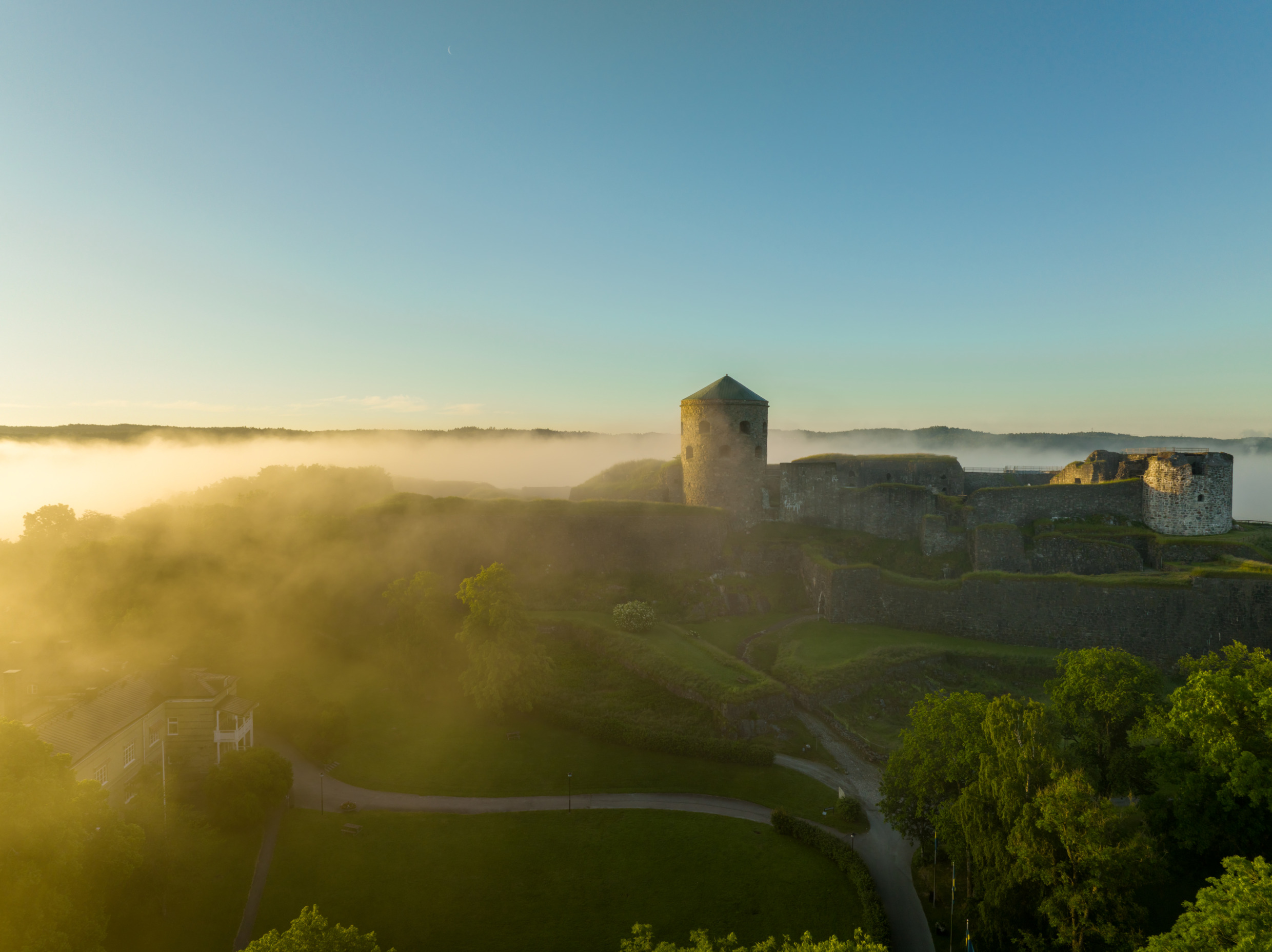 A drone view over Bohus Fortress surrounded by fog and green trees  on a sunny day with clear blue sky.