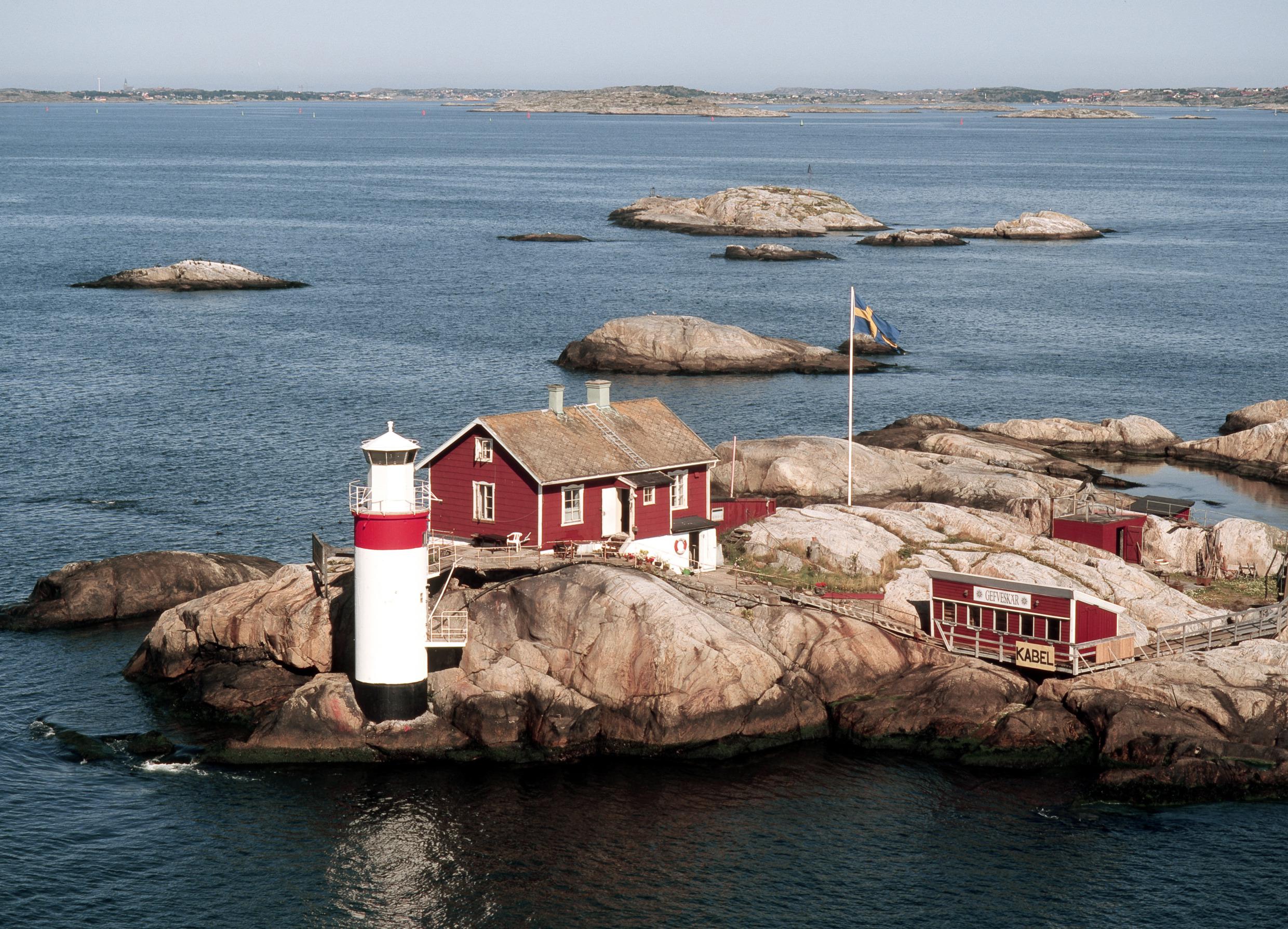 Ein rotes Holzhaus, ein weißer Leuchtturm und ein Fahnenmast mit der schwedischen Flagge auf einer Insel im Schärengarten.