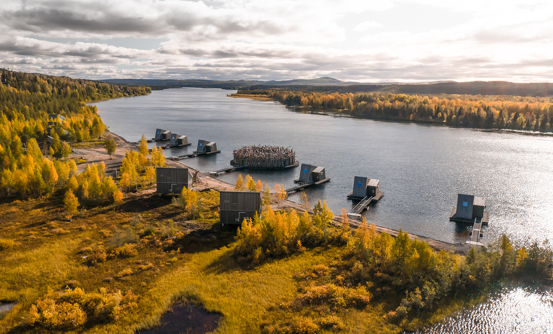 Aerial view of Arctic Bath in autumn, with cabins on land and floating on the river, surrounded by golden-leaved forest.