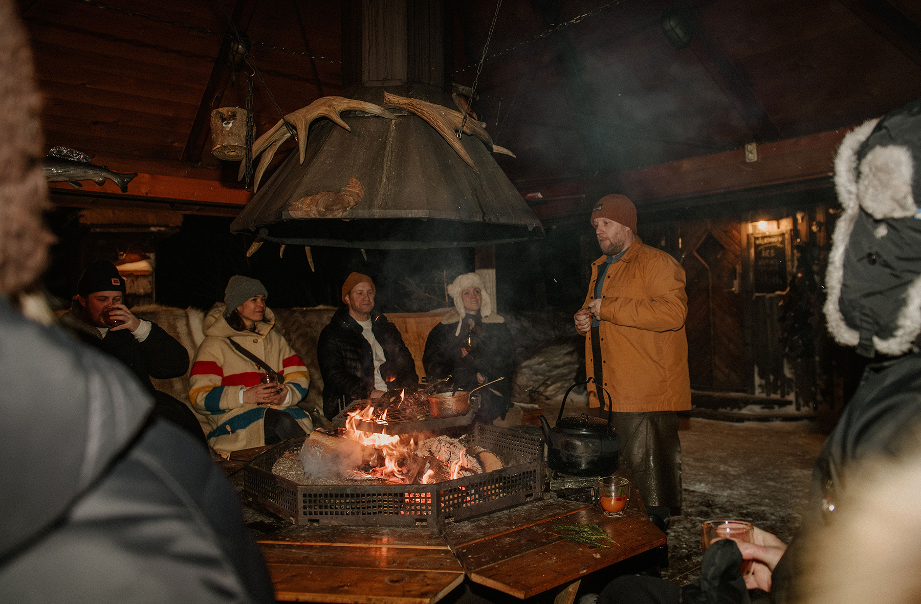 A group of people sitting around an open fire outdoors at night, cooking food and socialising in a snowy setting.