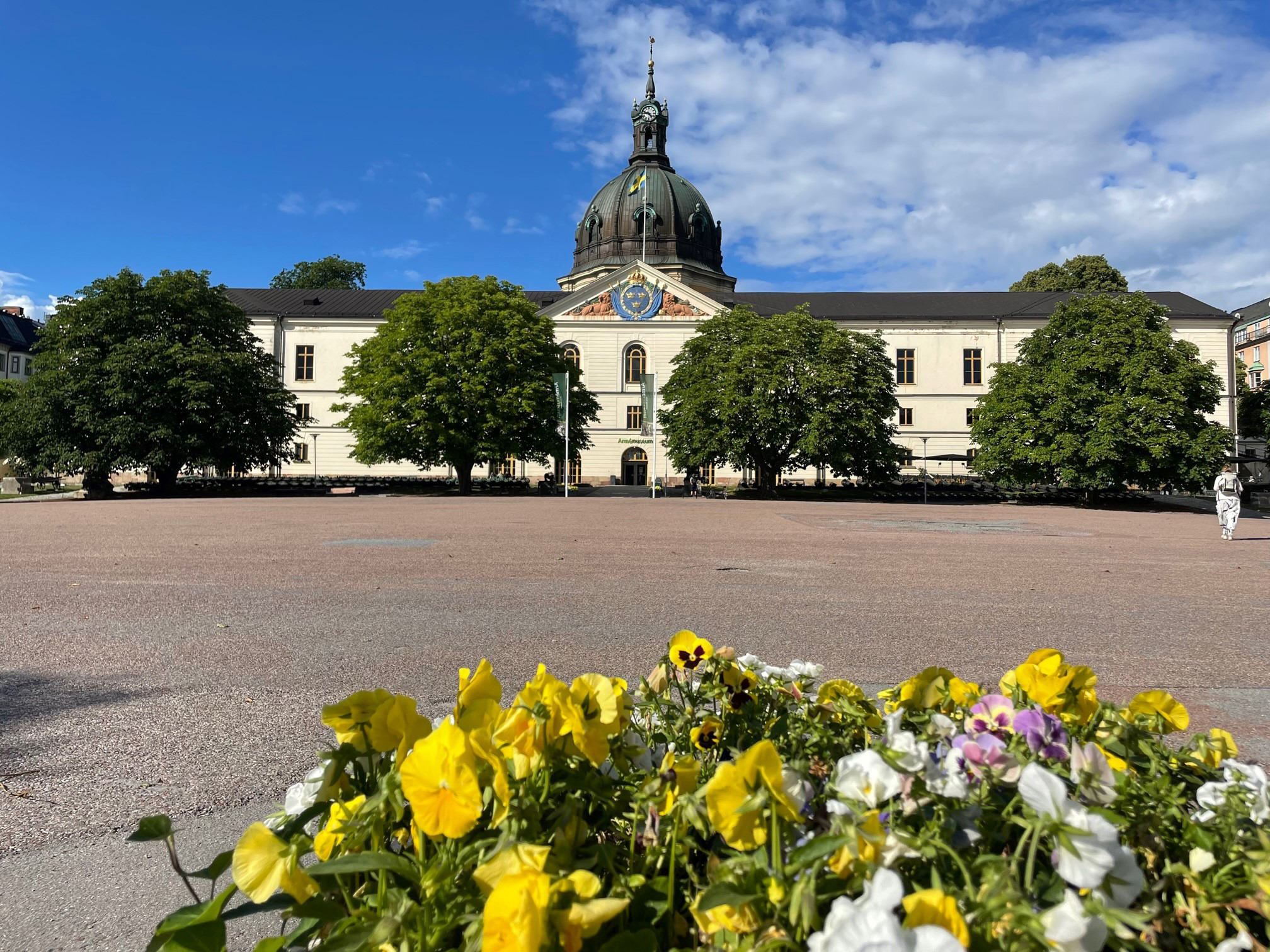 Buitenkant van het Legermuseum in Stockholm op een zonnige dag, met een bloembed op de voorgrond en het koepeldak dat boven de bomen uitsteekt.