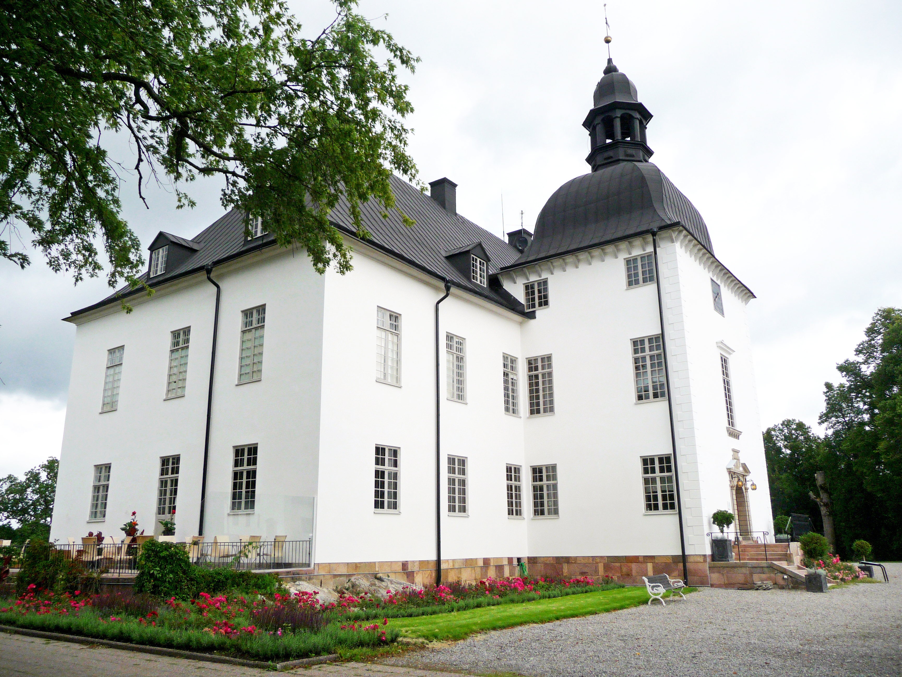 White baroque-style building of Årsta Castle in Haninge, Sweden, with a black domed tower and surrounding gardens in summer bloom.