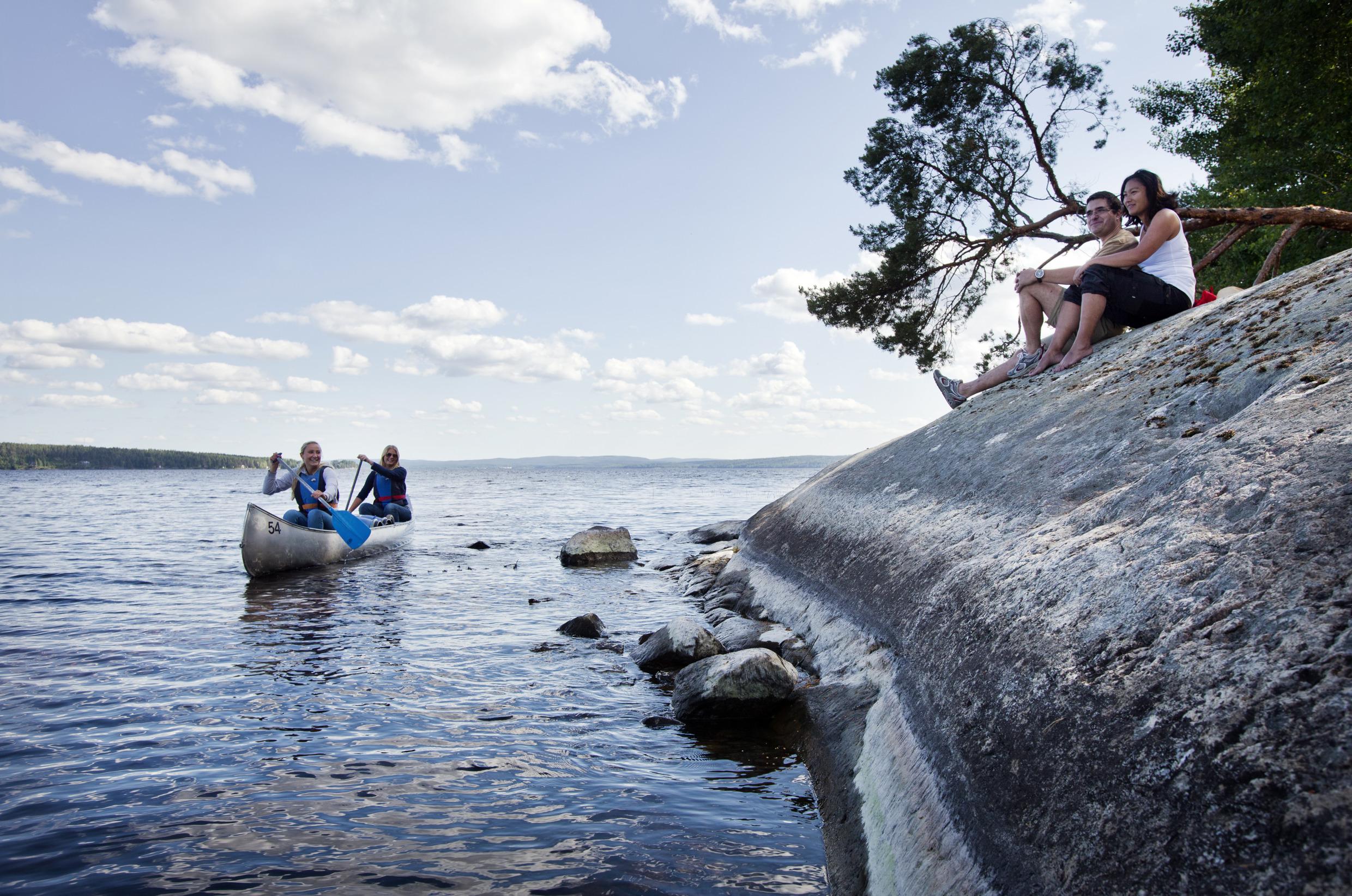 Een man en een vrouw zitten op een klif bij een meer terwijl twee vrouwen in een kano peddelen.
