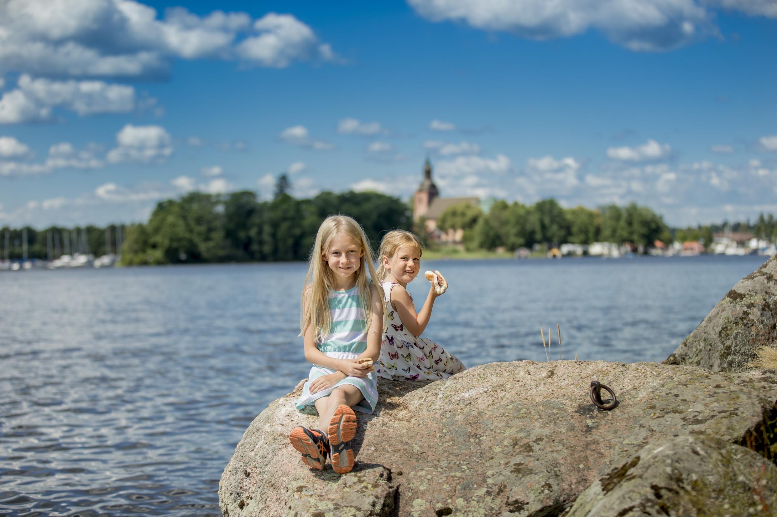 Two children, girls, sits on a cliff next to the water during summer.