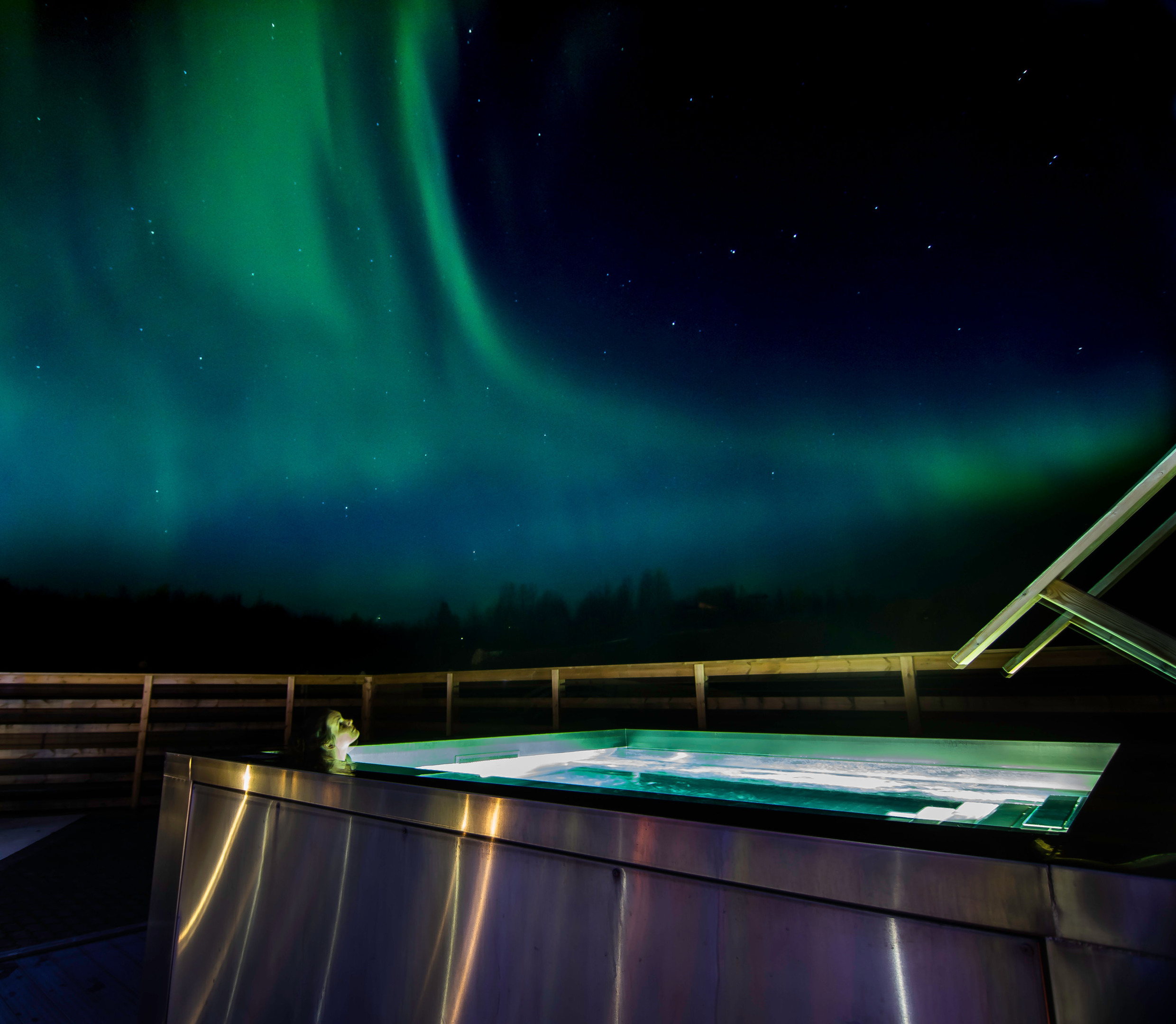 Eine Person sitzt in einem Whirlpool. Am Nachthimmel tanzen Nordlichter.